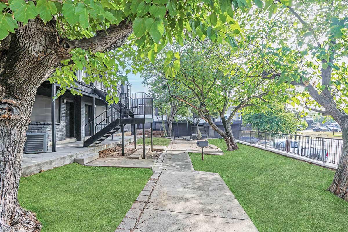A landscaped outdoor area featuring several trees, grassy paths, and a concrete walkway. There's a staircase leading to a multi-level building in the background. Grills are situated near the trees, creating a welcoming space for gatherings. The scene is bright and inviting, with ample sunlight filtering through the leaves.