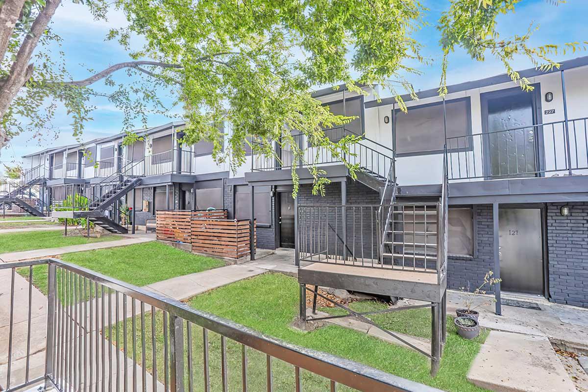 A view of a two-story apartment complex featuring multiple units with black metal railings and doors. The landscaping includes a grassy area and a tree, providing a green touch to the exterior. The building's facade is a mix of dark colors, and there are wooden privacy screens on the lower levels.