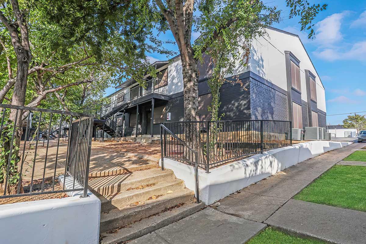A modern multi-unit building with a mix of dark and light-colored exteriors. The structure features a staircase leading to upper levels, surrounded by trees and landscaping. A landscaped area with grass is visible in the foreground, along with a concrete sidewalk.