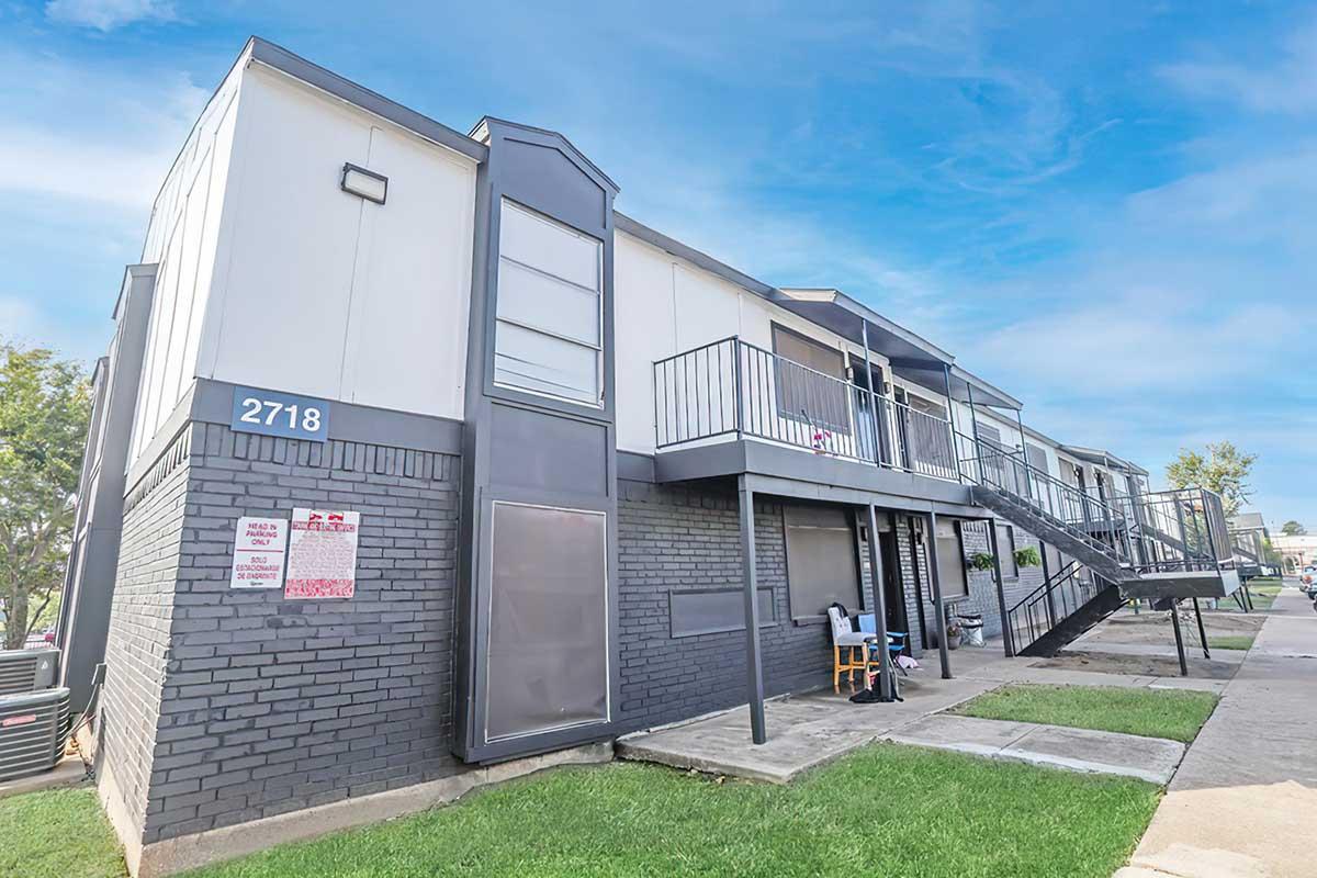 Two-story apartment building with a modern design featuring multiple units, a staircase leading to the upper level, and a small grassy area in front. The building has a mix of dark and light-colored walls, balconies, and a sign visible near the entrance. The sky is clear with soft clouds.