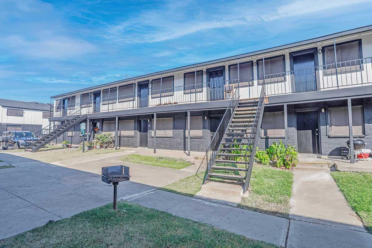 A two-story apartment building with a simple architectural design, featuring several balconies and stairs leading to the upper level. The building is surrounded by grass and has a small barbecue grill area in the foreground. The sky is partly cloudy, adding a bright atmosphere to the scene.