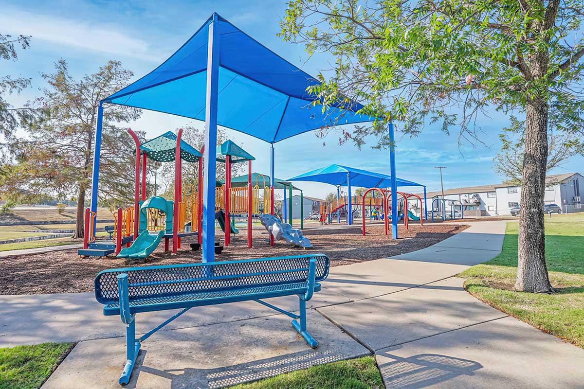 A colorful playground with various play structures, including slides and climbing areas, shaded by large blue canopies. In the foreground, there is a blue bench. The area is well-maintained, surrounded by grass and trees, with a pathway leading through the playground. The sky is clear and sunny.