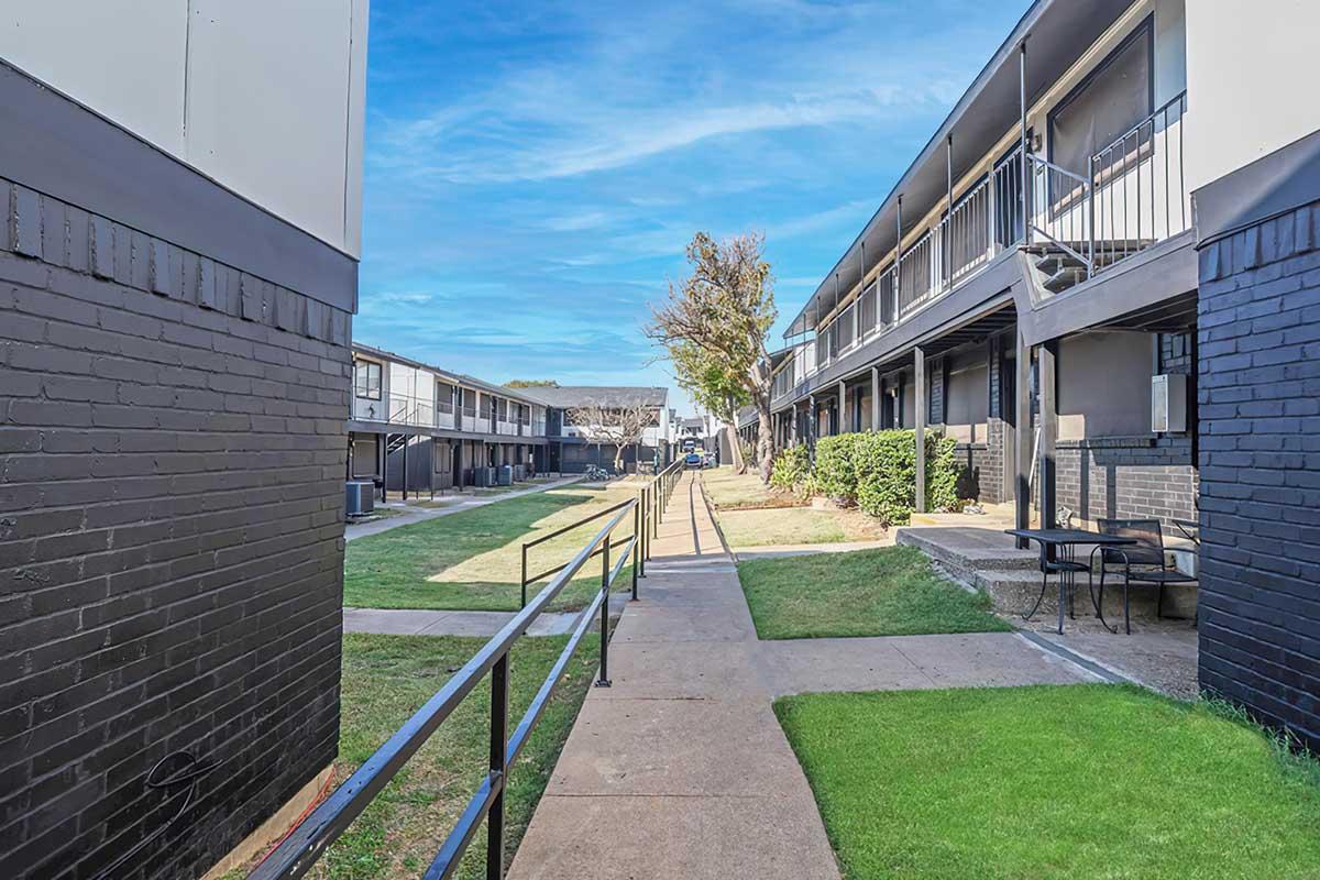 View of a residential apartment complex featuring two buildings on either side of a landscaped pathway. The pathway is flanked by green grass and small trees, with outdoor seating visible. The sky is bright and clear, creating a welcoming atmosphere.