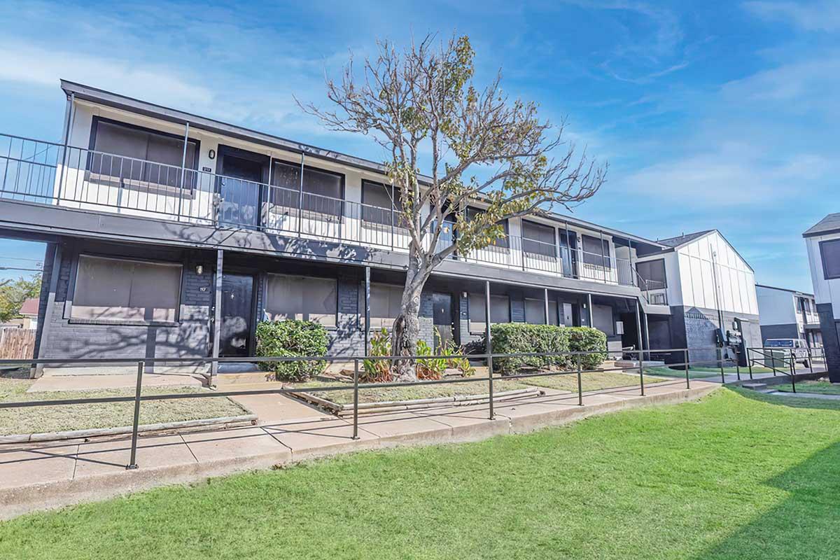 A two-story apartment building with a balcony on the upper floor. There is a small tree and green grass in the foreground, along with a paved pathway leading to the entrance. The sky is clear and blue, complementing the modern design of the building.