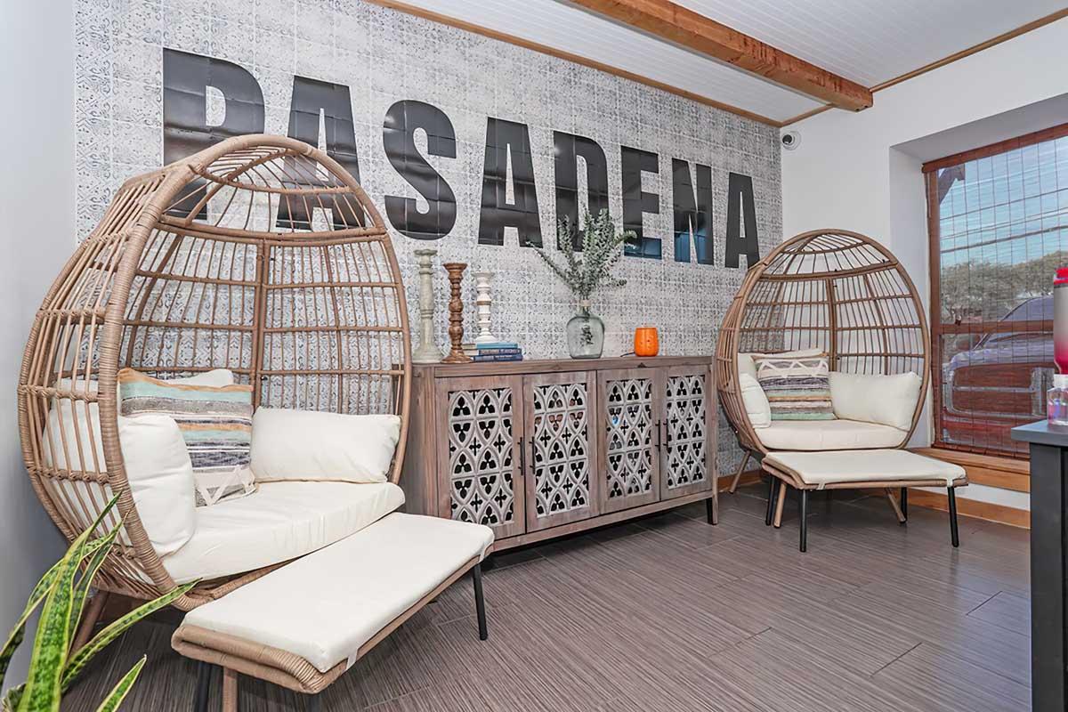 A cozy interior space featuring two stylish woven chairs facing each other, a decorative wooden sideboard in the center, and a large wall displaying the word "PASADENA." There are touches of greenery and decorative items that add warmth and character to the room. Natural light streams in through a window.