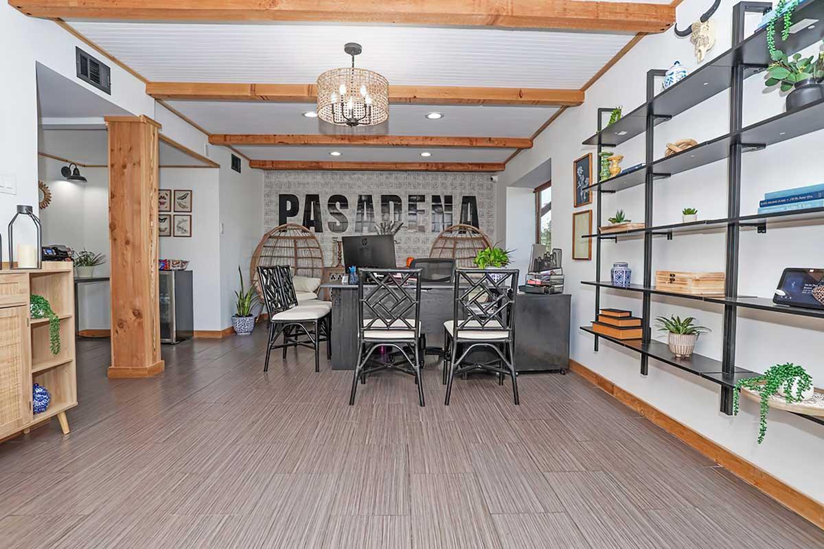A modern office reception area featuring a welcoming atmosphere. The space includes a large sign that reads "PASADENA," a front desk with stylish chairs, wooden beams, and decorative shelves displaying plants and books. Natural light brightens the area, creating an inviting environment for visitors.