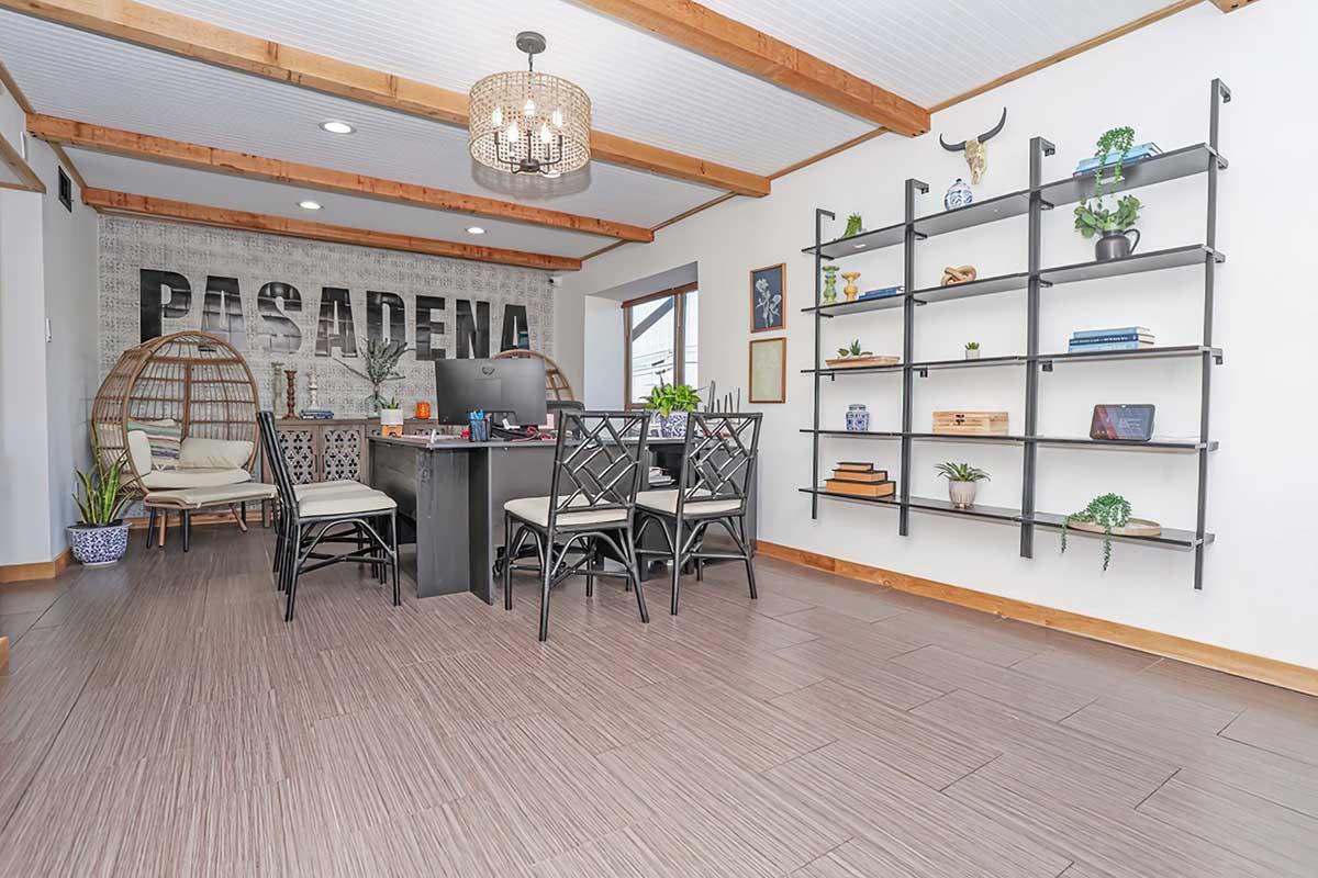 A modern interior space featuring a dining area with a black table and four chairs, decorative plants, and shelving displaying books and decorative items. The wall displays the word "PASADENA," and wooden beams accent the ceiling, creating a trendy and inviting atmosphere.