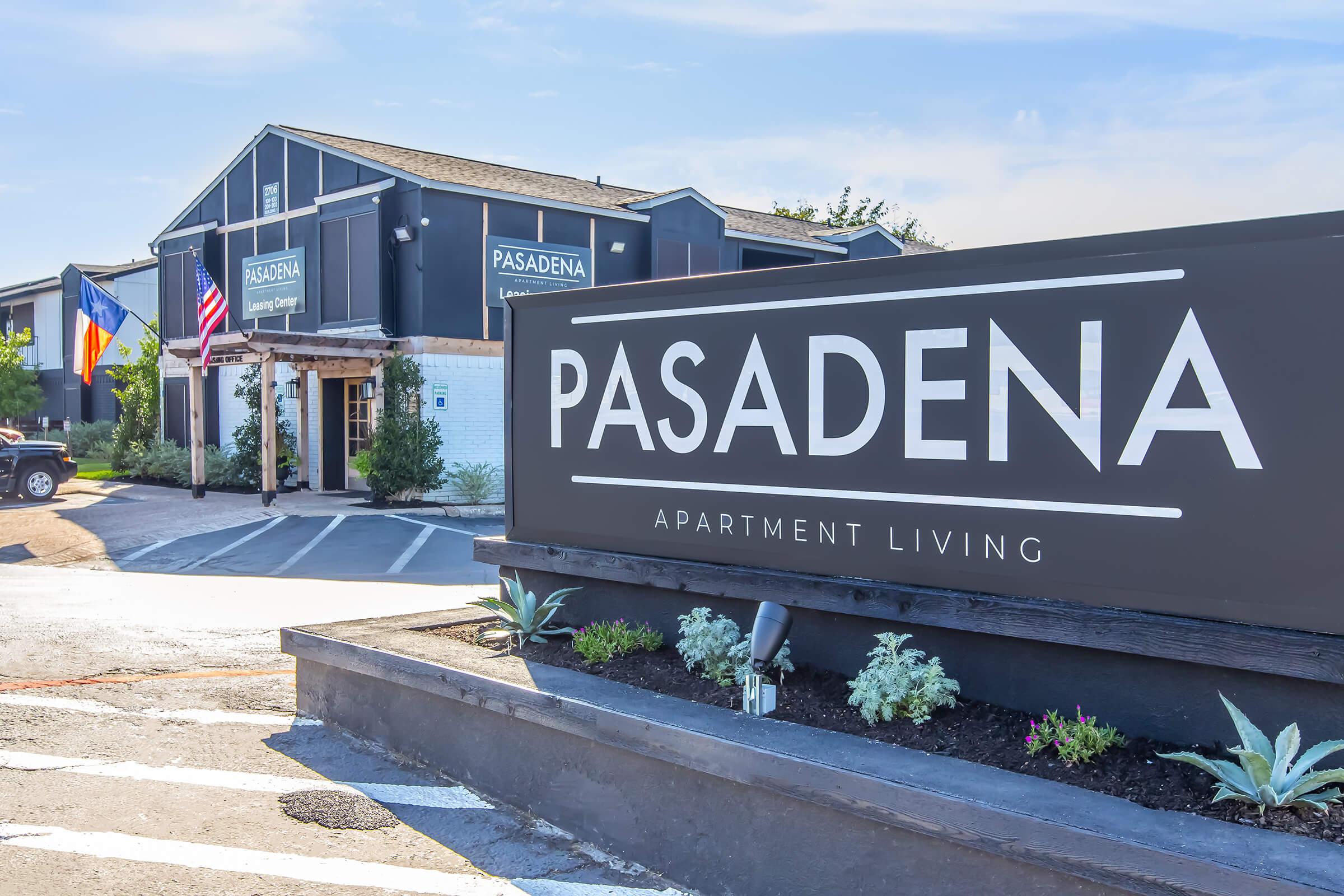 Sign for Pasadena Apartment Living, prominently displayed in front of a modern building. The building features a dark exterior with lush greenery, and flags representing the United States and another country are visible. Parking area in the foreground with neatly landscaped plants.