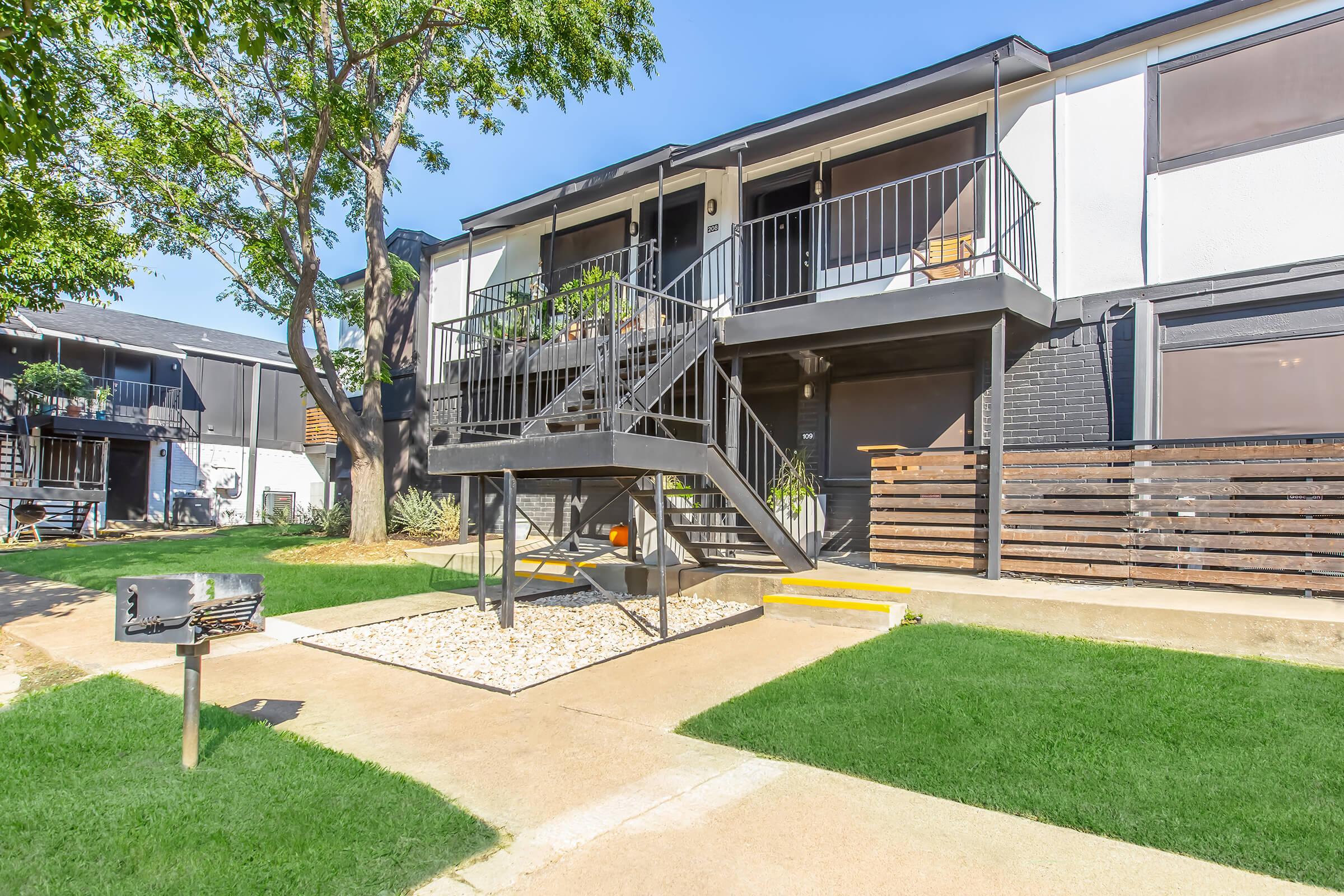 Two-story apartment building with a black metal staircase leading to the second floor. Grass and gravel landscaping surround the entrance, which is marked by a small mailbox. The building features a modern design with large windows and a balcony. Trees provide shade in the area.