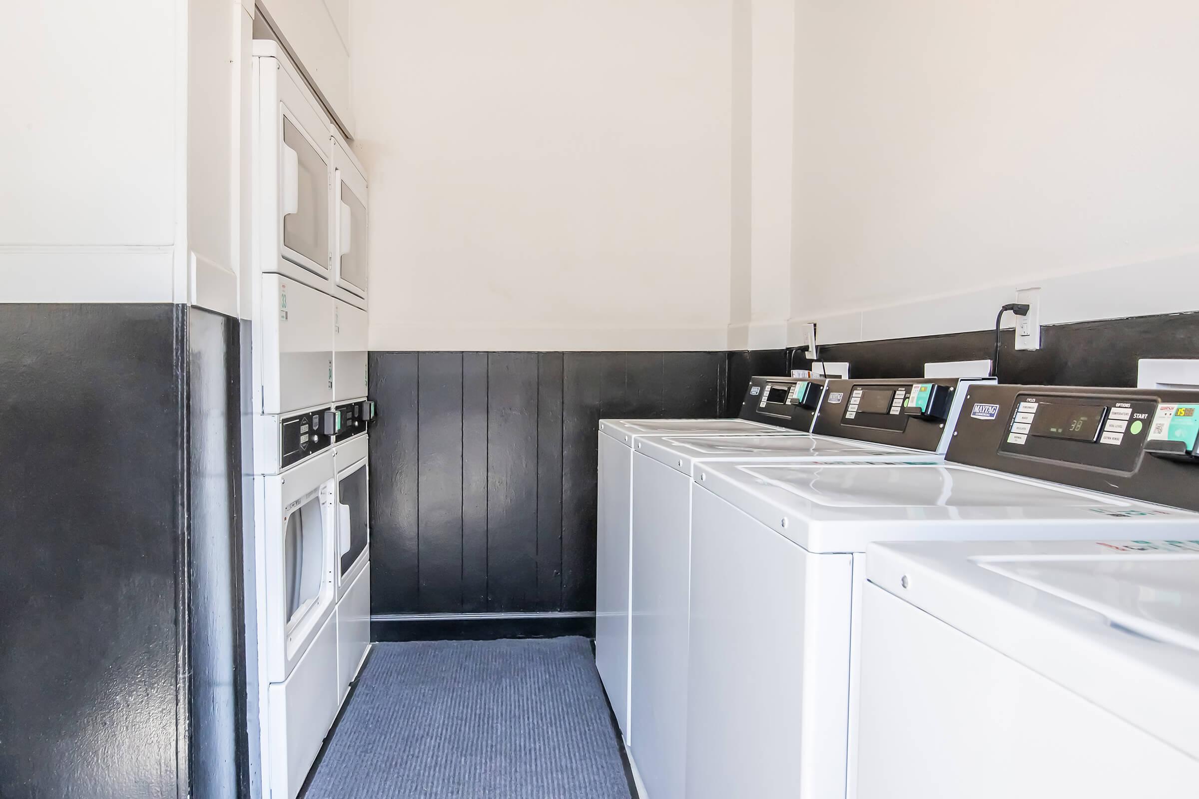 A clean and organized laundromat featuring multiple washing machines and dryers. The machines are lined up along the wall in a bright space with light-colored walls and a black accent wall. A carpet runner covers the floor, creating a neat appearance in the area.