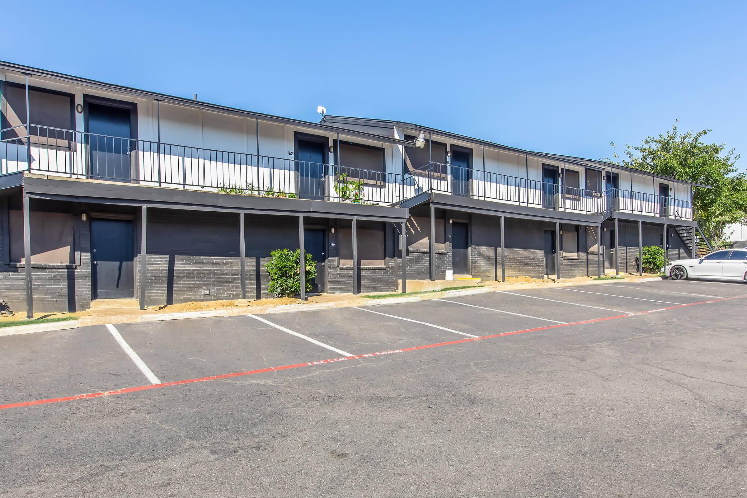 A two-story motel with multiple rooms, featuring balconies and outdoor stairs. The building is painted in dark colors and is surrounded by small shrubs. In the foreground, there are several empty parking spaces on a paved lot under a clear blue sky.