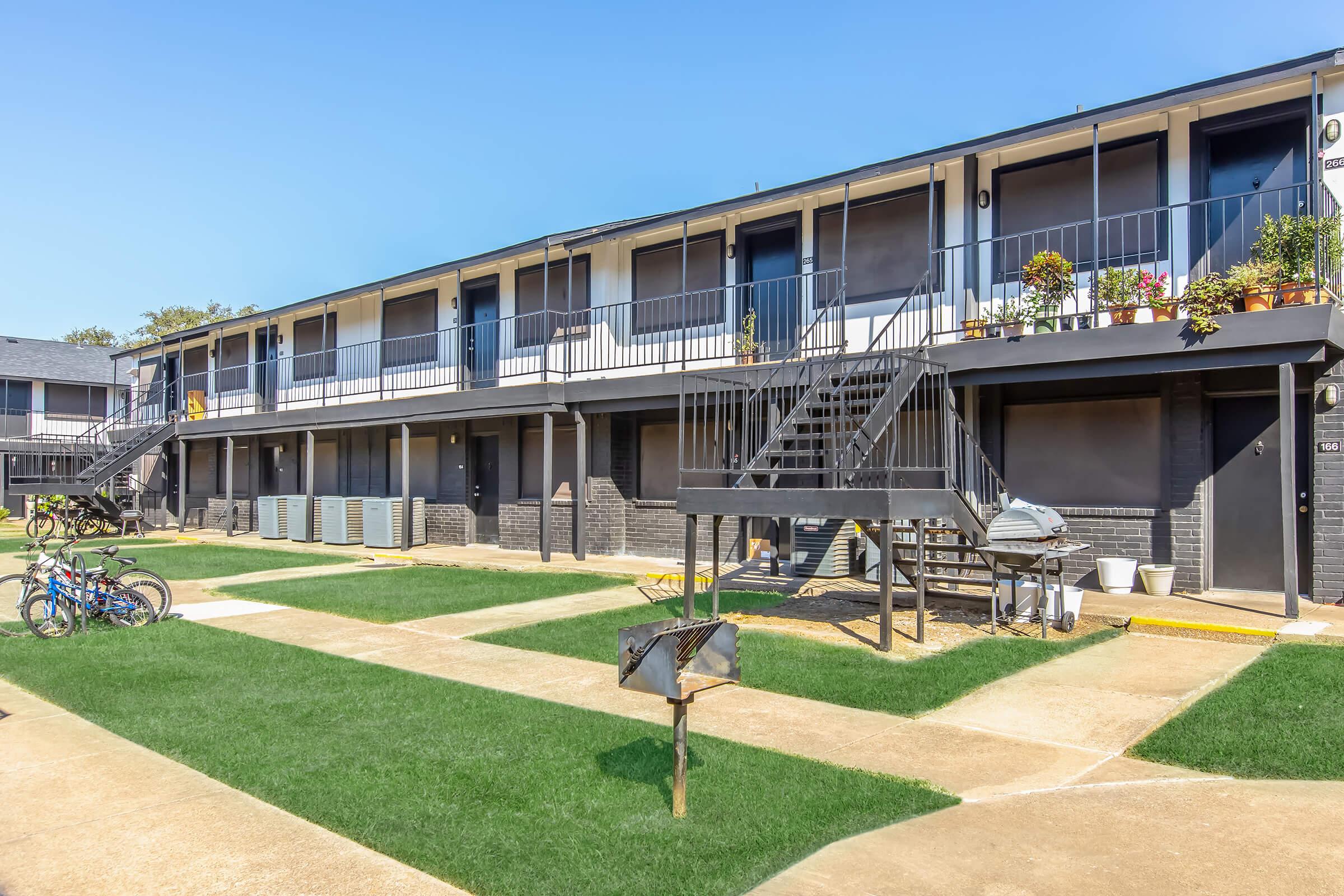 A view of a two-story apartment complex featuring multiple units with balconies. The courtyard has green grass, bike racks, and outdoor seating. Staircases lead to the upper level, while air conditioning units are visible along the building's exterior. Clear blue sky in the background.