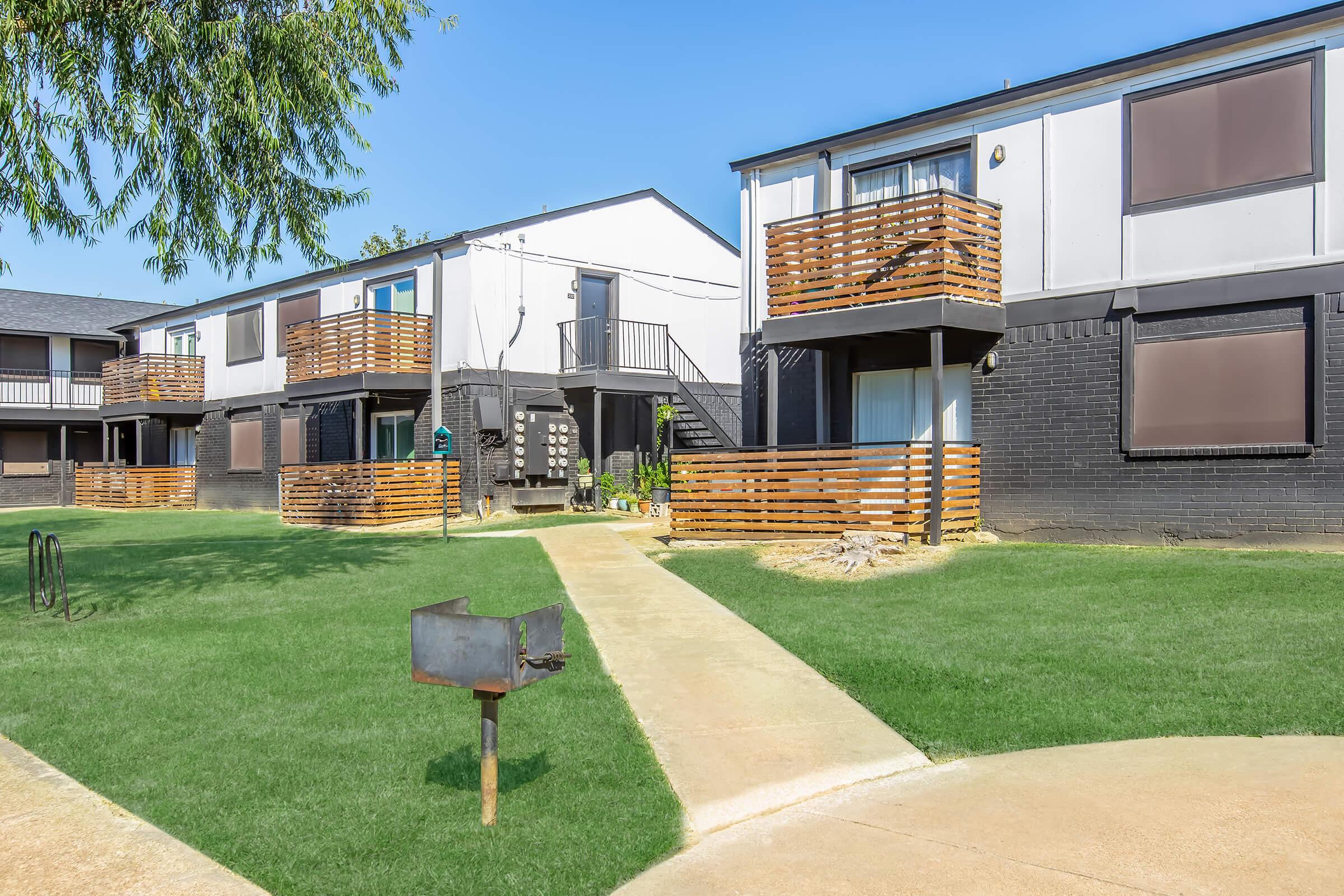 A view of a multi-unit apartment complex with two buildings connected by a pathway. Each building features balconies with wooden railings. The grassy area is well-maintained, and there's a mailbox near the pathway. Clear blue sky visible in the background.