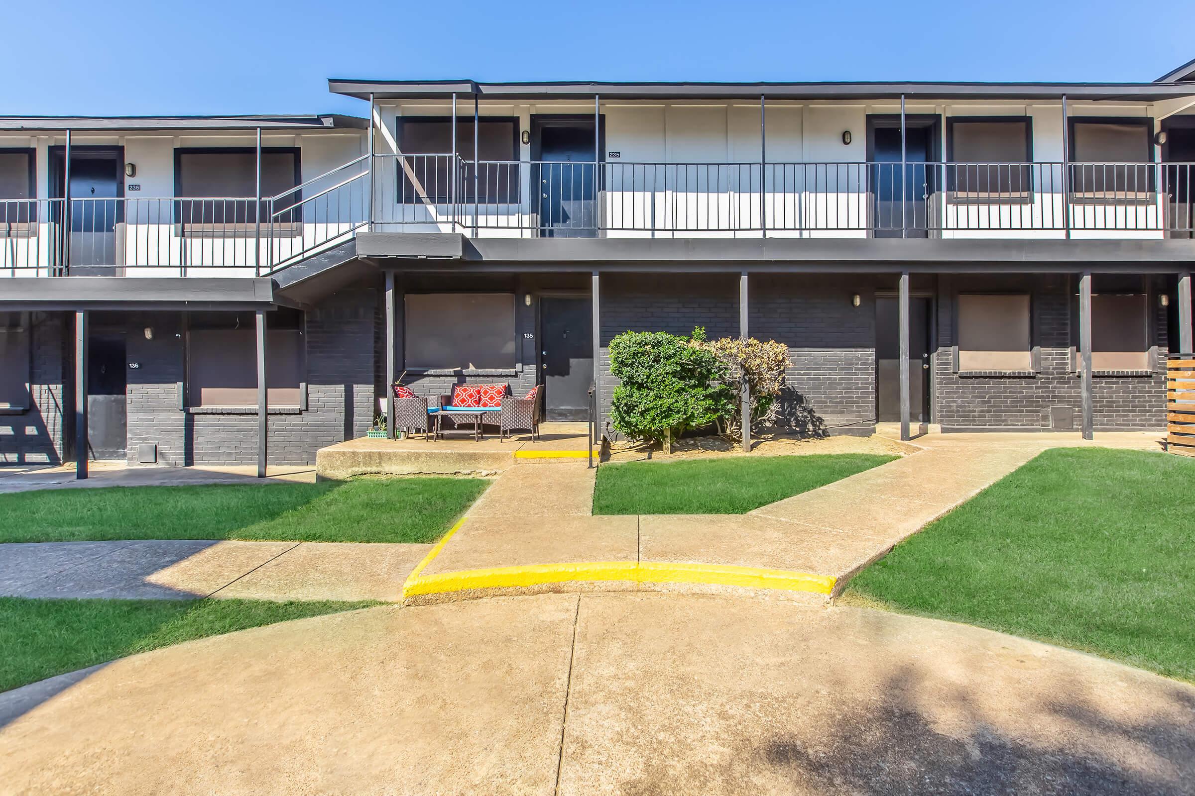 A view of a two-story apartment complex with a modern design. The building features several balconies and a pathway lined with green grass. A small seating area with red cushions is visible in front, and there are concrete walkways leading to the entrances of the units.