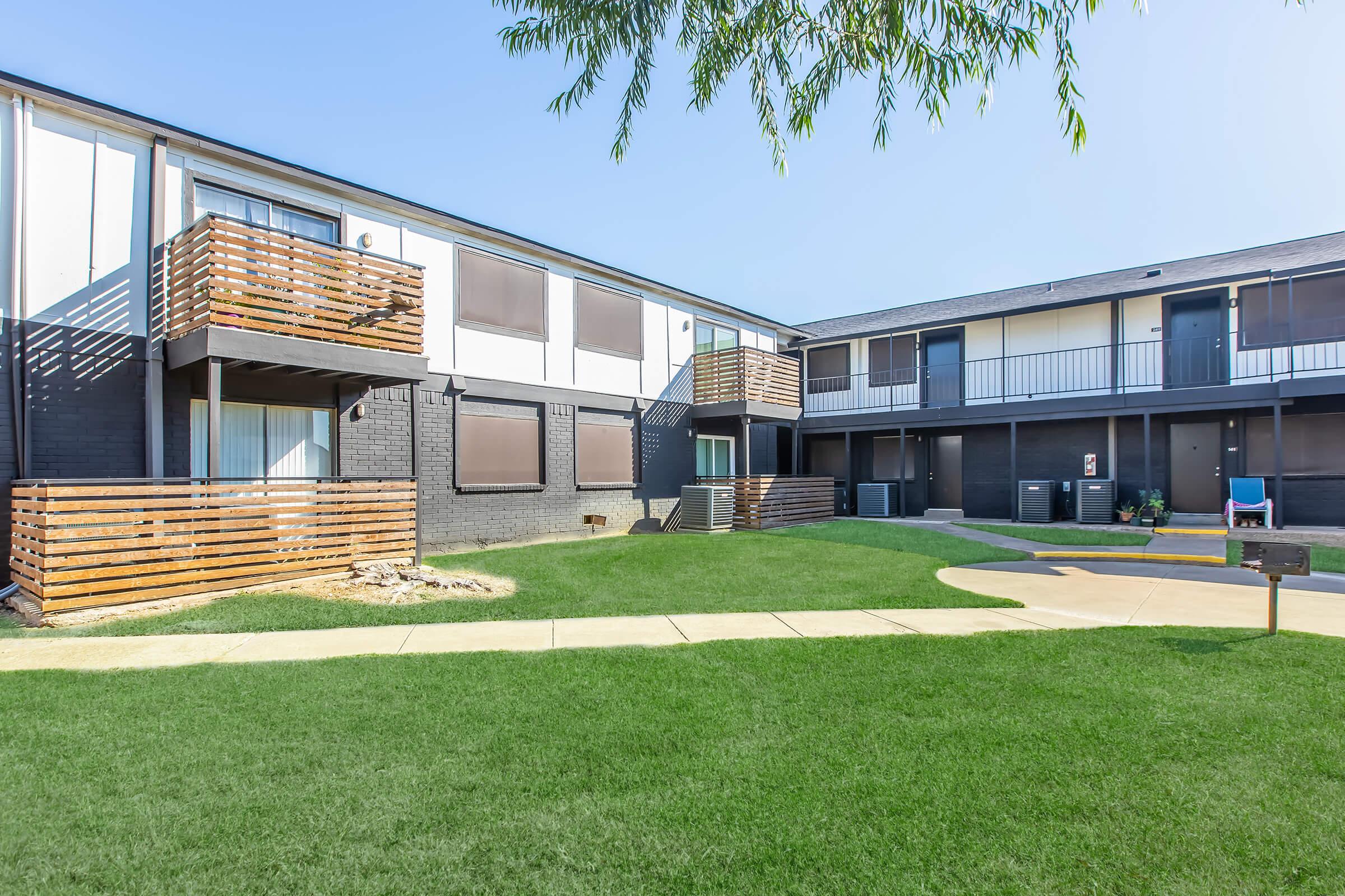 View of a modern apartment complex featuring two buildings with a grassy courtyard in the foreground. The buildings have balconies with wooden railings and are painted in light and dark colors. The sky is clear and the area is well-maintained, creating a welcoming outdoor space.