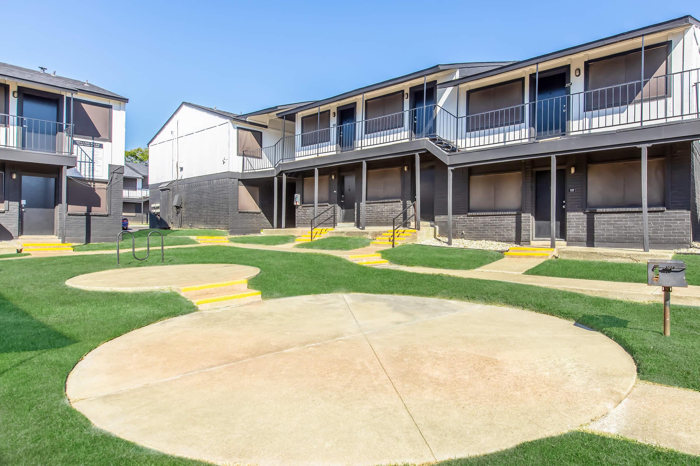 A view of a multi-unit apartment complex featuring two buildings with balconies. The well-maintained lawn includes circular concrete pathways and brightly painted yellow steps leading to the entrances. A clear blue sky is visible above, enhancing the inviting atmosphere of the property.