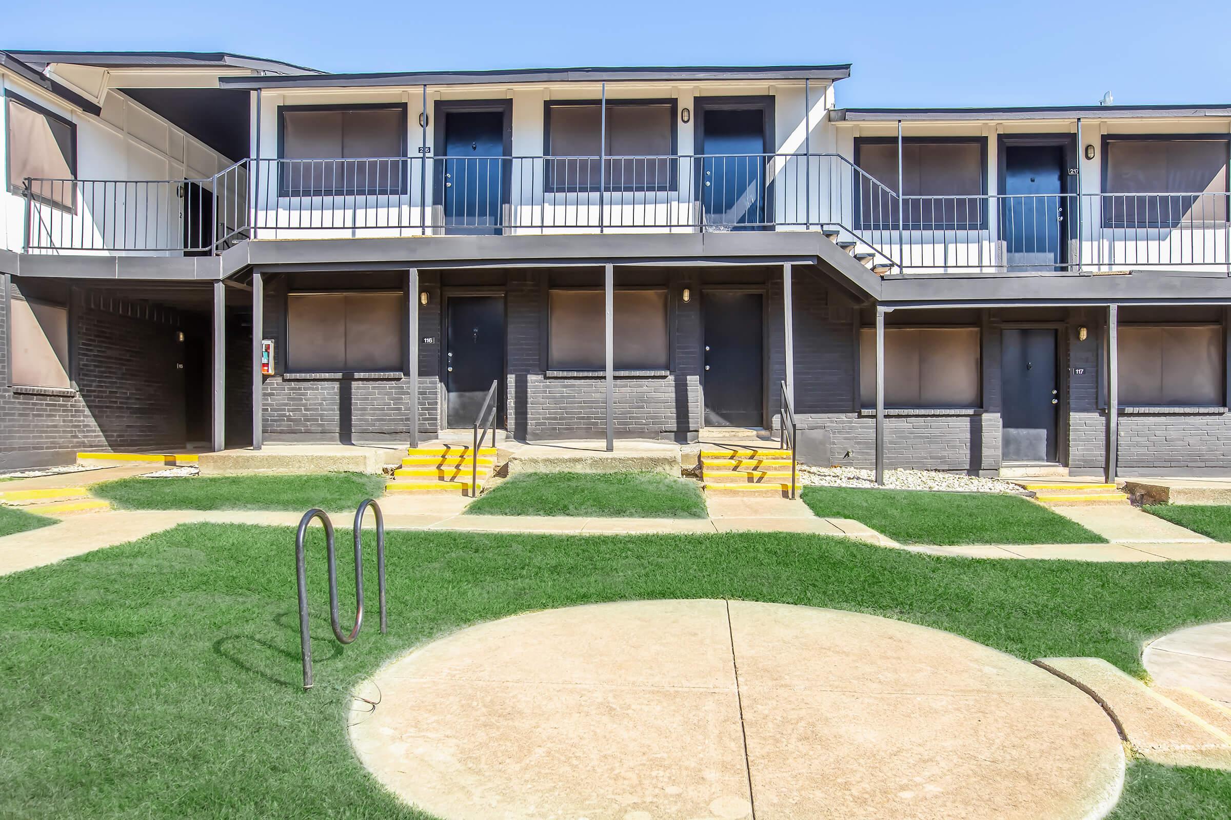 A two-story residential building featuring multiple entrances with black doors and white railings. The building is set against a bright blue sky and includes a pathway leading to a grassy area. There are steps leading up to each door, and a circular concrete pad is visible in the foreground.