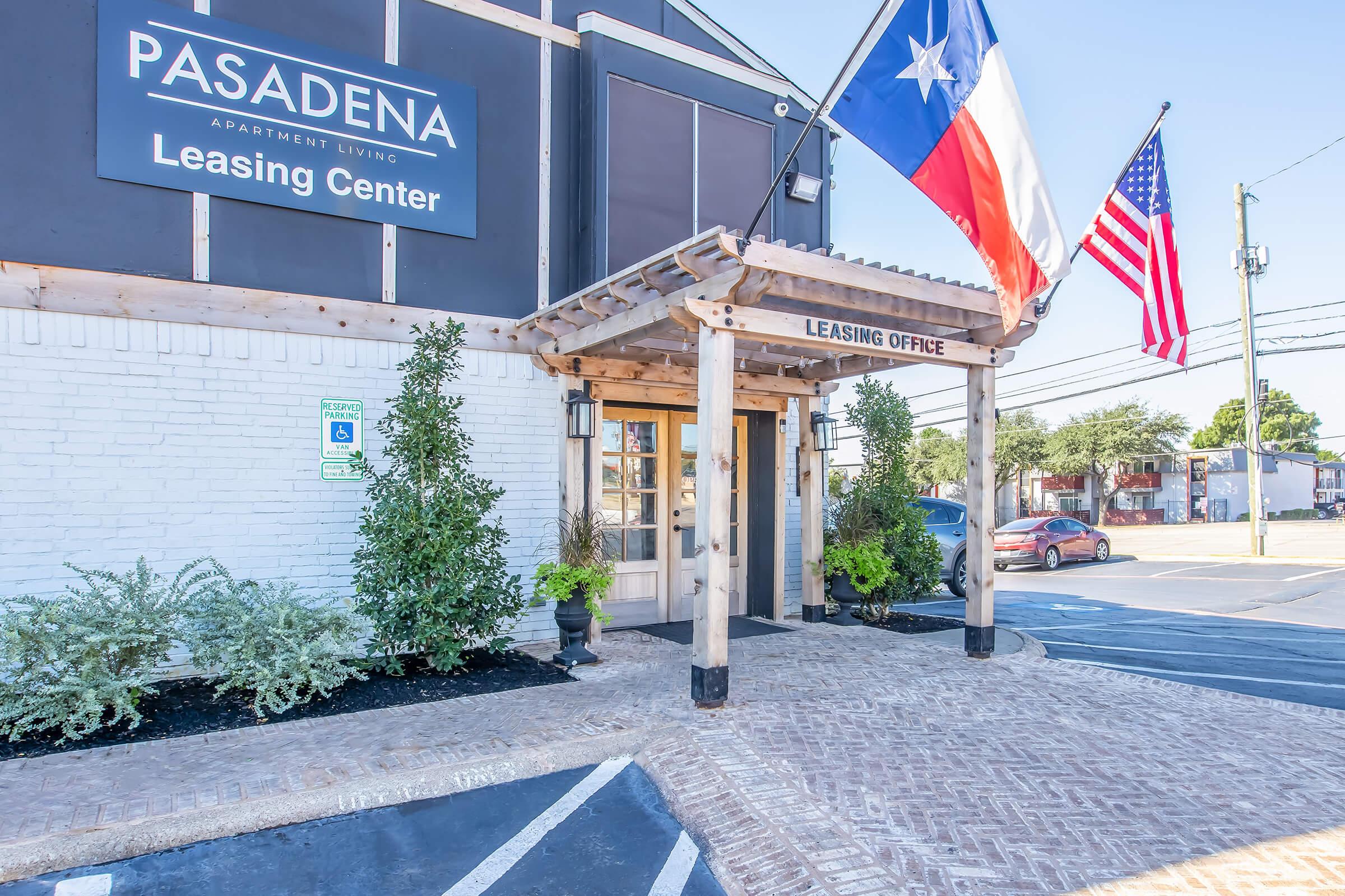 Entrance to the Pasadena Apartment Living Leasing Center, featuring a wooden pergola with flags of Texas and the United States. The building has a modern exterior with brick pathways and landscaped greenery. A sign indicates the leasing office and there's a parking area visible nearby.