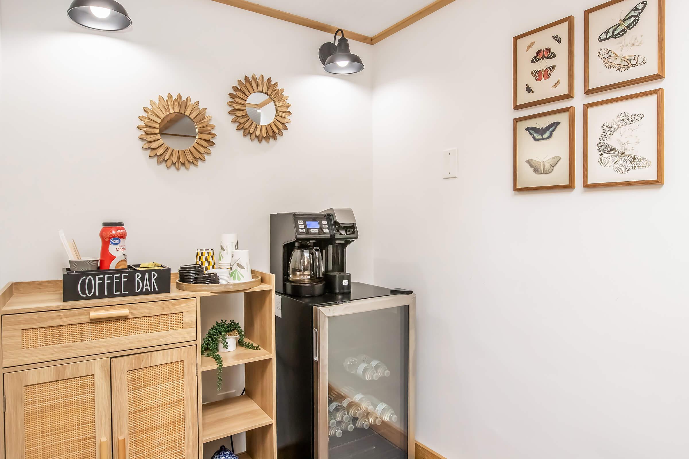 A cozy coffee corner featuring a coffee maker, a cabinet labeled "Coffee Bar," and a small fridge. The wall is adorned with framed butterfly and moth illustrations, complemented by two decorative mirrors. Soft lighting creates a warm and inviting atmosphere.