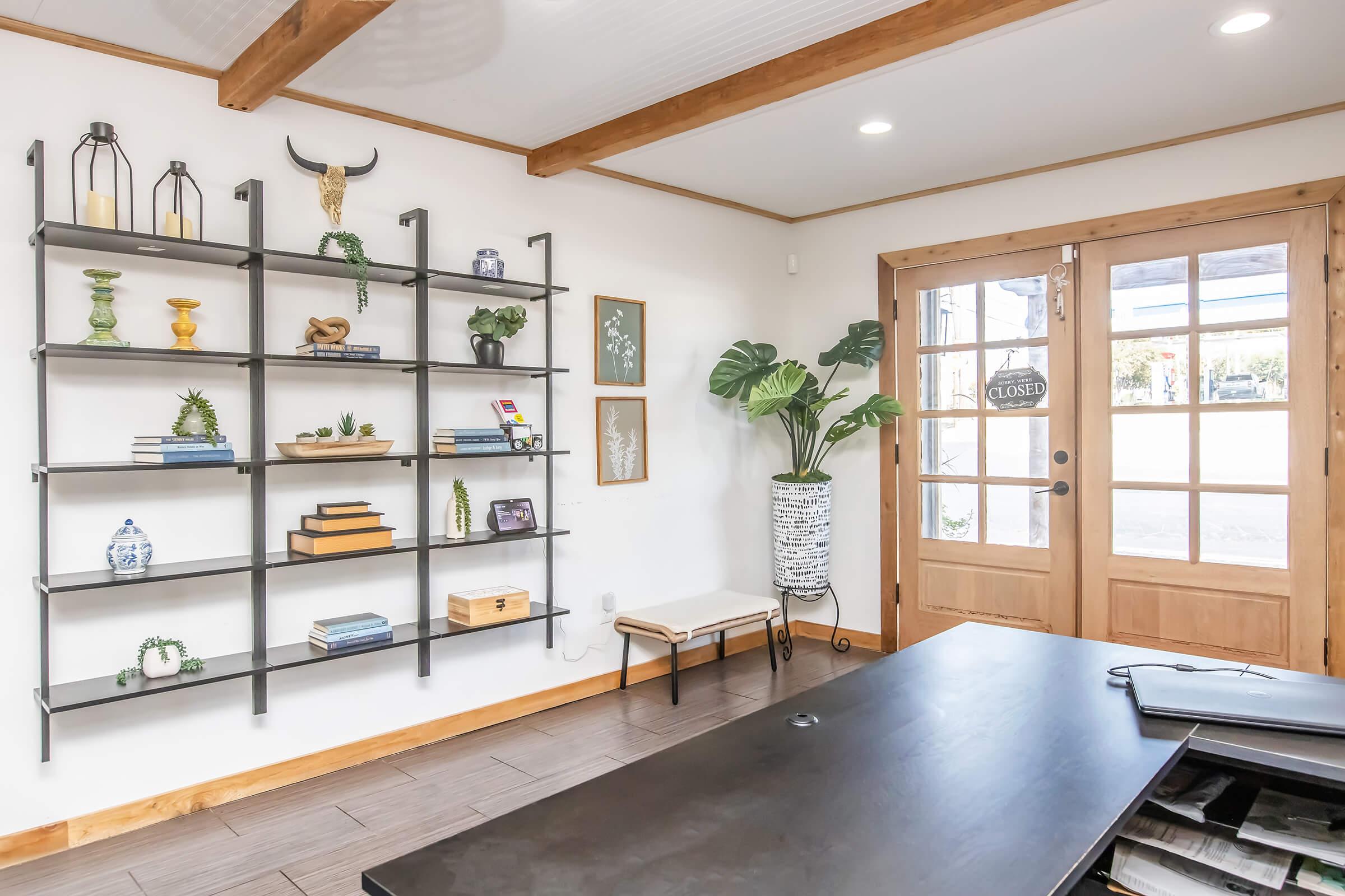 A modern interior space featuring a black metal shelf with various decorative items, including plants, books, and sculptures. There's a wooden door with glass panels showing a "CLOSED" sign, and potted plants are placed in the corner. The flooring is a light wood, creating a warm atmosphere.