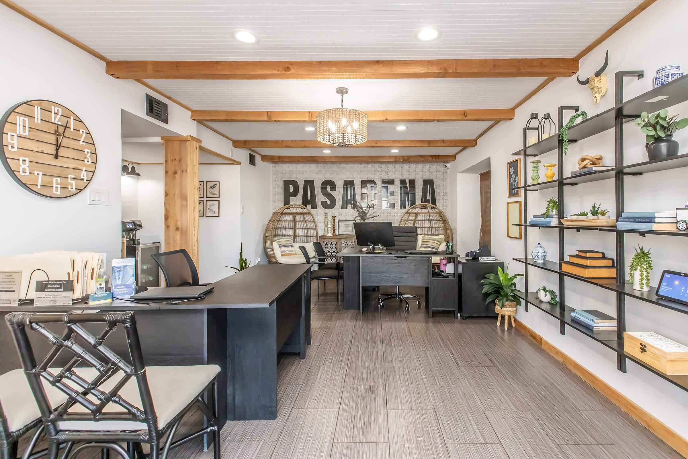 Modern office reception with wooden beams, a large decorative wall featuring "PASADENA," and stylish shelving displaying books and plants. A black desk is centered with a computer and office chairs, alongside a round wall clock and two large decorative baskets. Brightly lit and inviting atmosphere.