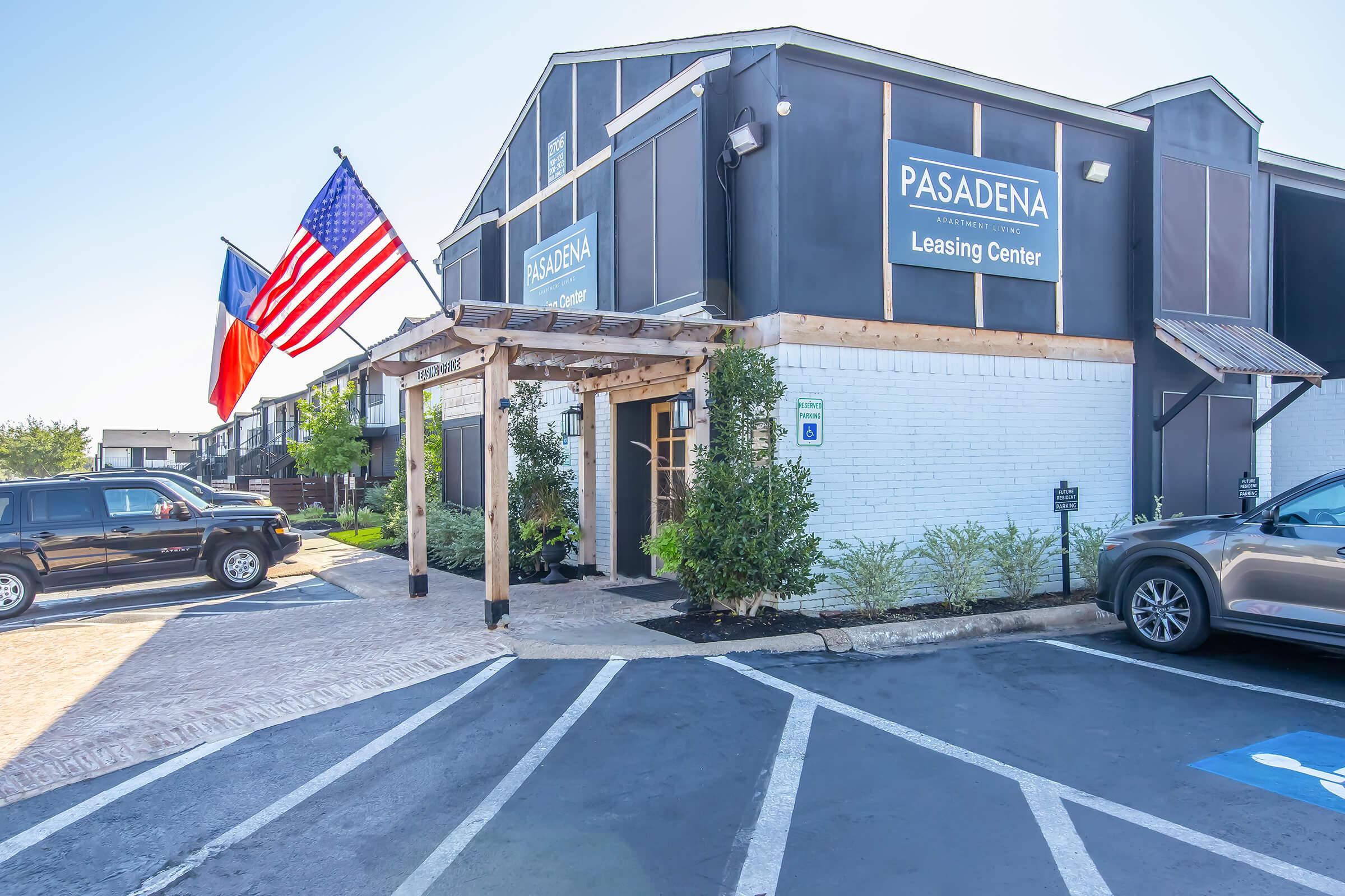 Leasing center building with a porch, featuring American and Texas flags. There is a well-maintained parking lot with a few parked cars and greenery around the entrance. The building has a modern design with signs showing it is the Pasadena Leasing Center. Clear blue sky in the background.