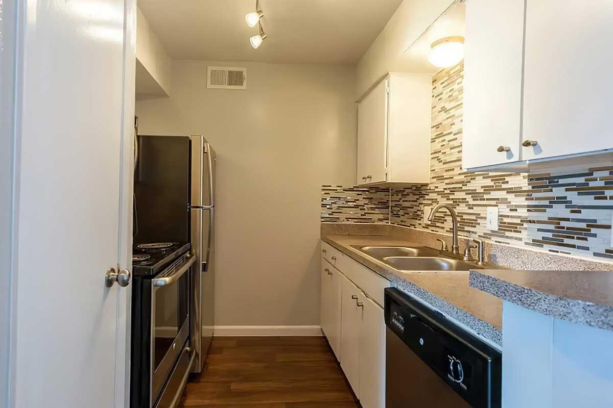 A modern kitchen featuring stainless steel appliances, including a fridge and stove. The cabinetry is white with a subtle granite countertop. A decorative tile backsplash adds texture, while warm wooden flooring completes the contemporary look. Good lighting highlights the space.