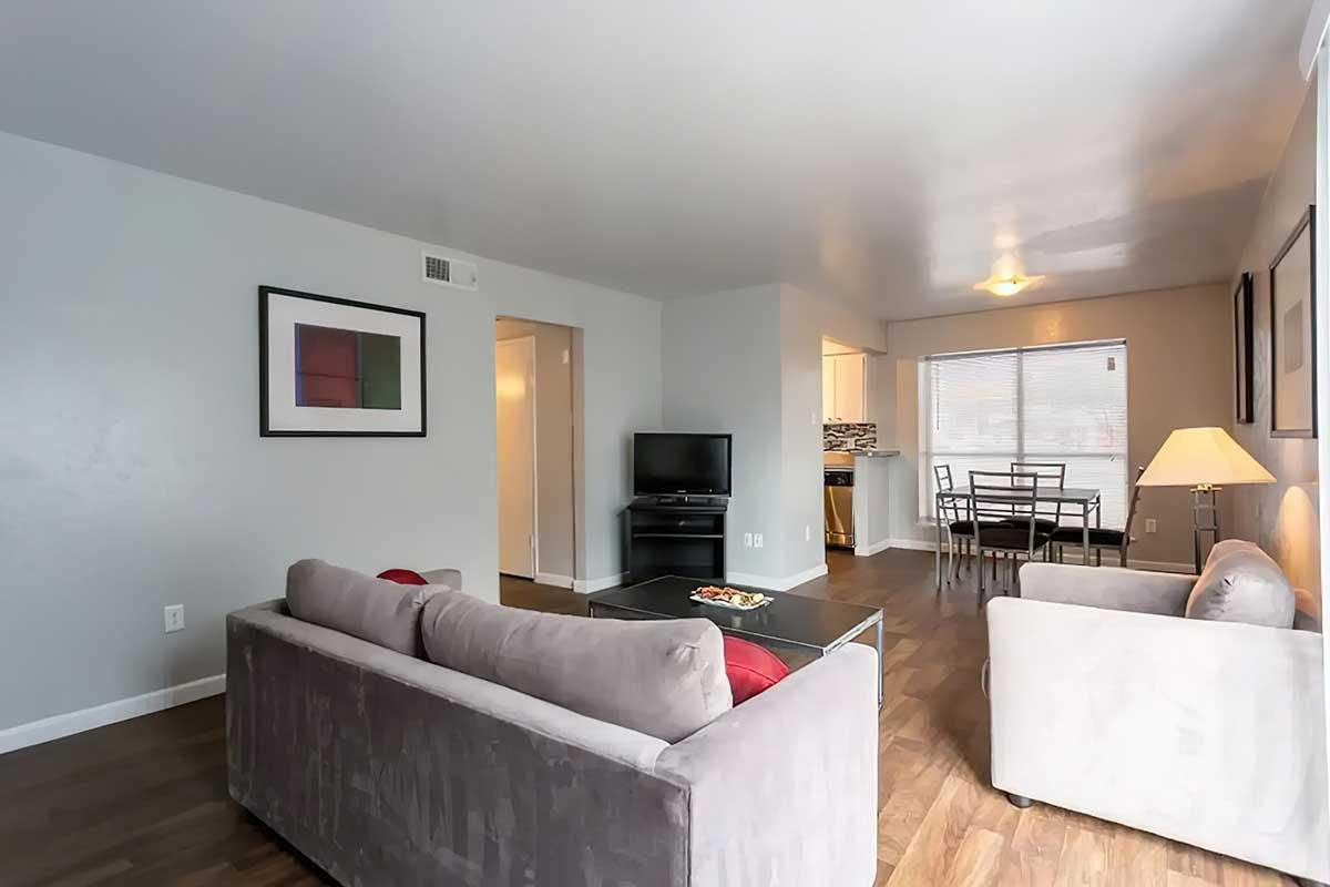 A modern living room featuring a gray sofa with red accents, a dark coffee table, and a flat-screen TV. There is a dining area with a table and chairs, large windows allowing natural light, and minimalist wall art. The flooring is wood, enhancing the contemporary aesthetic of the space.