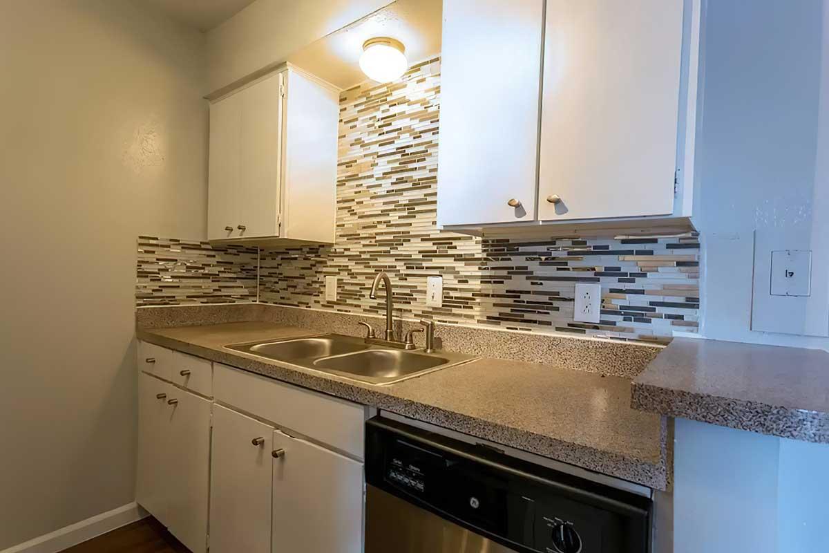 A modern kitchen featuring a double sink, granite countertop, and a stylish backsplash made of gray and white tiles. The cabinets are white, and there is a dishwasher underneath the countertop. The overall color scheme is light and minimalistic, creating a clean and inviting space.
