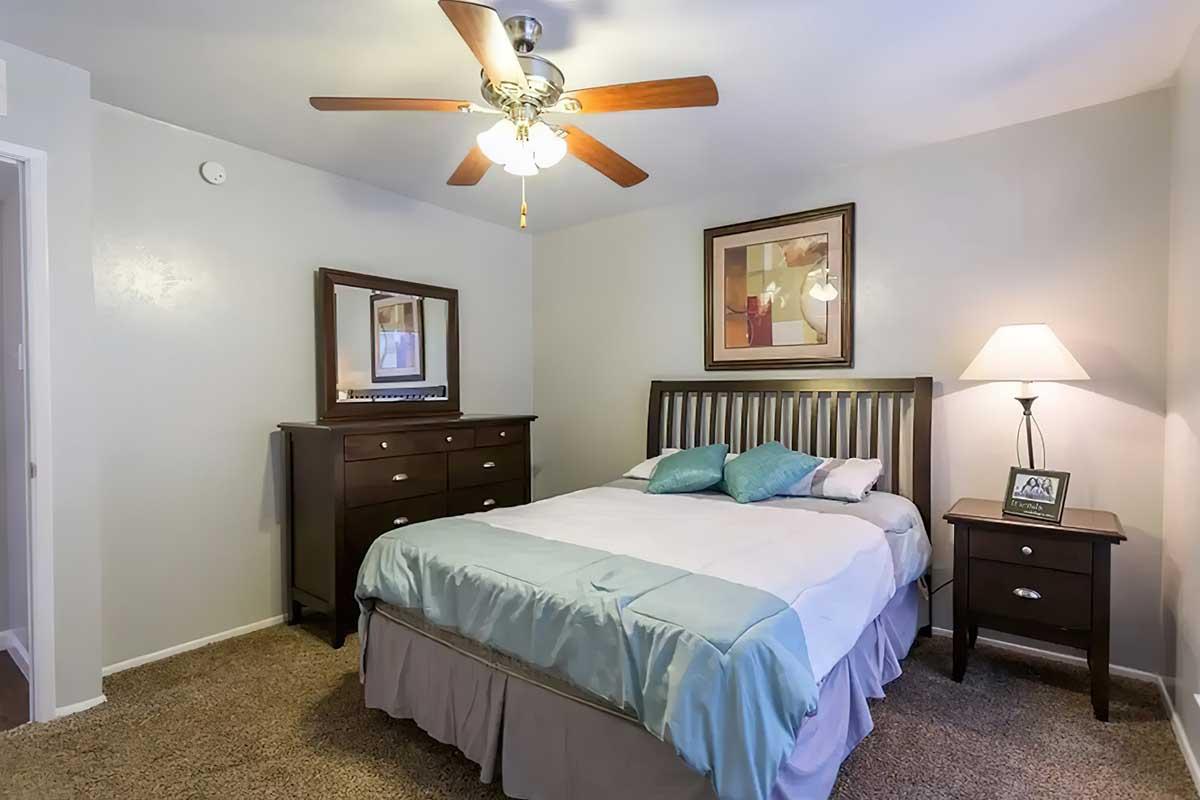 A cozy bedroom featuring a queen-sized bed with light blue bedding, a wooden headboard, and complementary nightstands. There is a ceiling fan above, a framed artwork on the wall, and a dresser with a mirror. Soft lighting from a lamp creates a warm and inviting atmosphere.