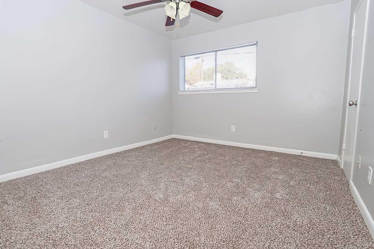 Empty room with light gray walls and carpeted flooring. A ceiling fan with wooden blades hangs from the ceiling. A single window provides natural light, and a white door is visible on the right side. The space is uncluttered and simple, ideal for personal decoration.