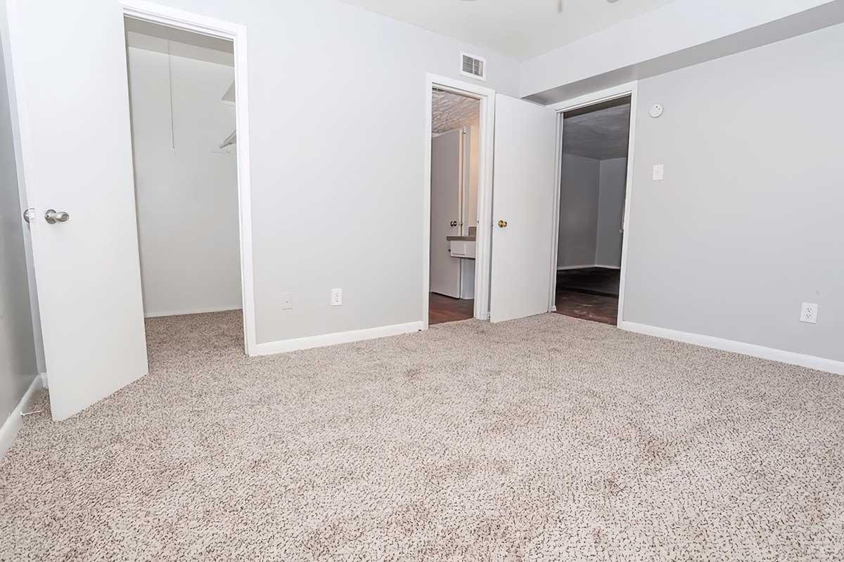 Spacious, empty room with gray walls and beige carpet. There are two open doors leading to other rooms, and no furniture is present. The natural light softly fills the space, creating an inviting atmosphere. Ideal for a fresh start or new decor ideas.