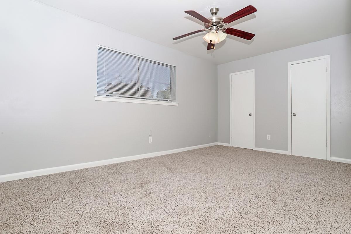A well-lit, empty room with light gray walls and beige carpet. It features a ceiling fan with wooden blades, a window with blinds, and two white doors on opposite walls. The room is spacious and clean, creating a welcoming atmosphere.