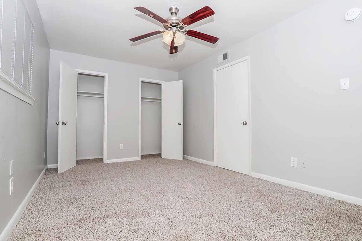 Empty room featuring neutral gray walls and carpet, with two closet doors visible on the left side. The ceiling has a red ceiling fan with three light fixtures, and there are two doors leading to other rooms. The space is well-lit and appears spacious.