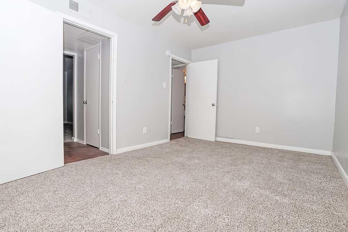 A spacious, empty room with light gray walls and a ceiling fan. The floor is covered with beige carpeting. There are two doorways leading to other rooms, with one doorway partially visible. The overall ambiance is bright and clean, suitable for various uses.