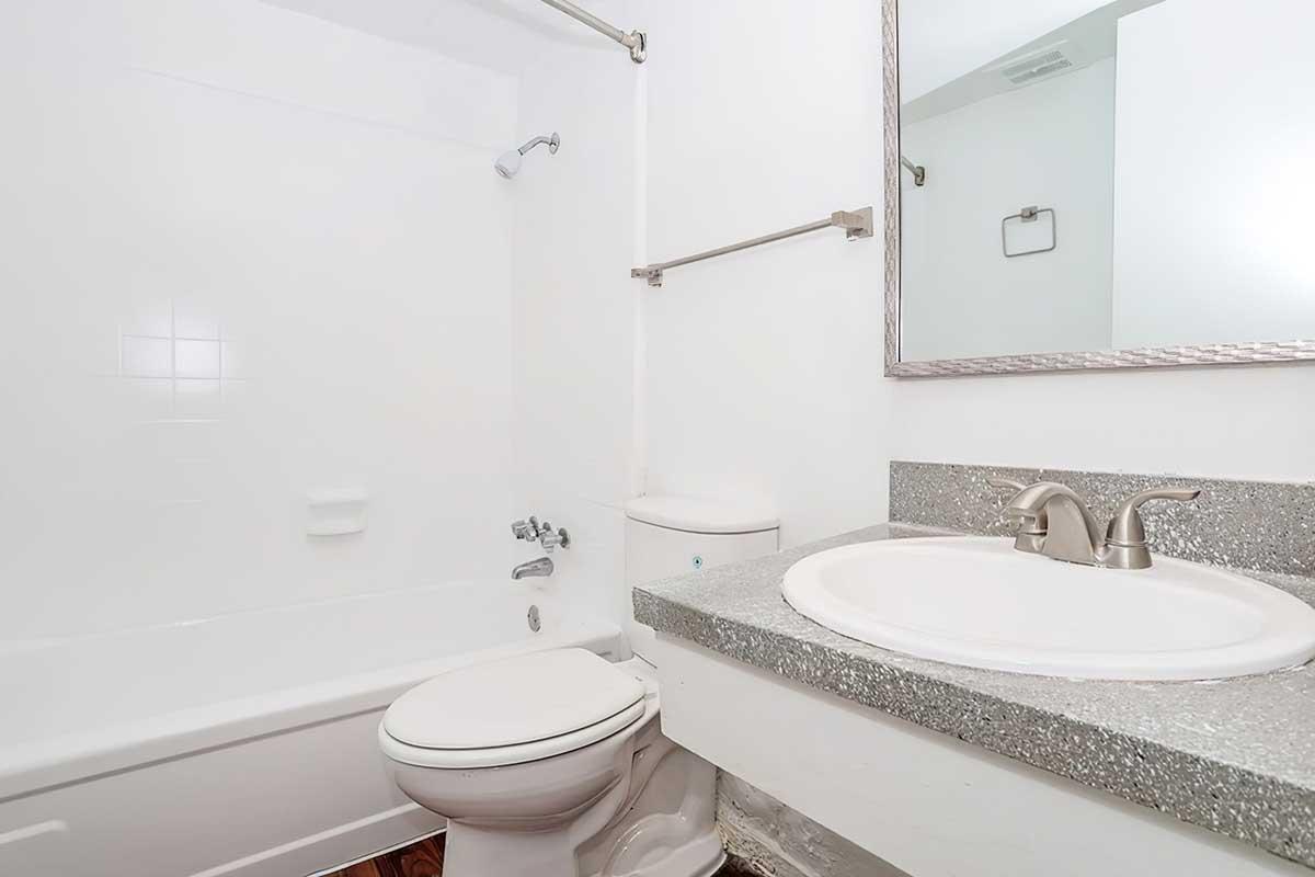 A clean and simple bathroom featuring a white bathtub, toilet, and sink with a granite countertop. The walls are painted white, and there is a mirror above the sink. The overall design is minimalist and bright, with a polished look.
