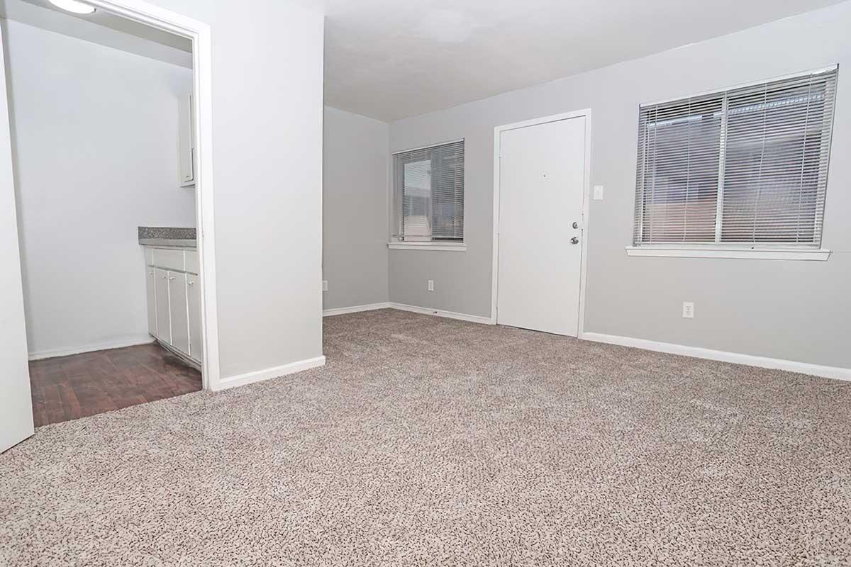 A spacious, empty room with light gray walls and a carpeted floor. On one side, there is a doorway leading to a small kitchen area, and on the opposite wall, a front door and two windows with blinds allow natural light to enter. Overall, the area looks clean and ready for furnishing.