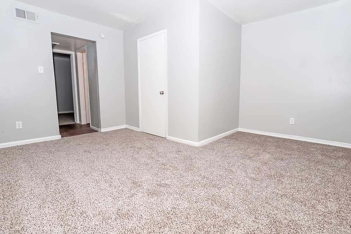 A spacious, empty room with light gray walls and beige carpeting. A closed white door is visible on the right, and there is an open doorway leading to another room in the background. Natural light brightens the area, creating a clean and inviting atmosphere.
