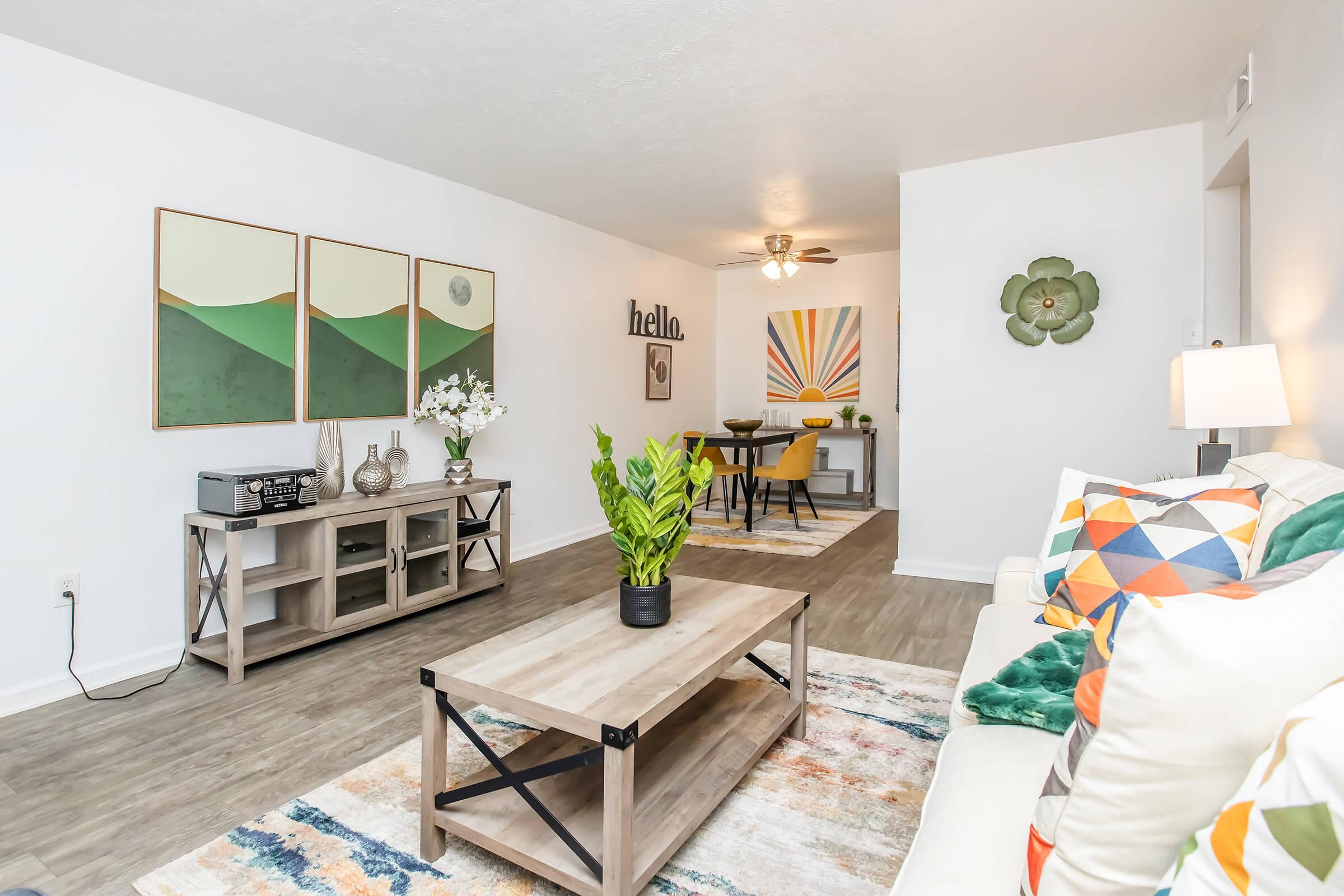 A modern living room featuring a beige sofa with colorful cushions, a wooden coffee table, and decorative plants. The wall showcases three framed artworks with abstract designs. A "hello" sign is displayed on the wall, and a dining area with a round table and chairs is visible in the background.