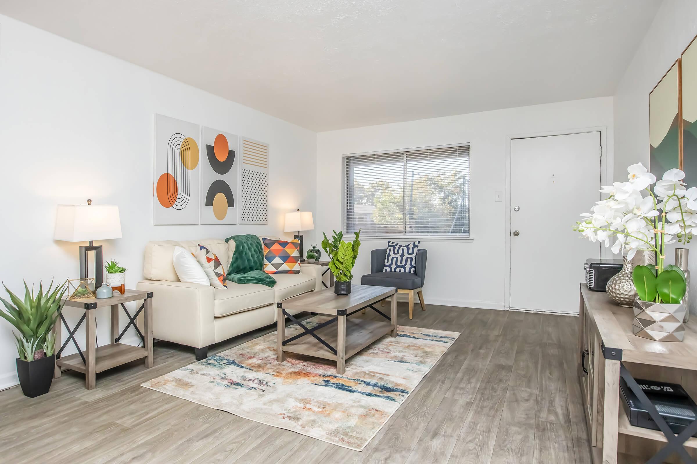 A cozy living room featuring a cream-colored sofa with colorful throw pillows, a glass coffee table, and decorative plants. There are two stylish chairs, a large artwork on the wall with abstract designs, and natural light coming through a window. The floor has a textured rug, enhancing the inviting atmosphere.