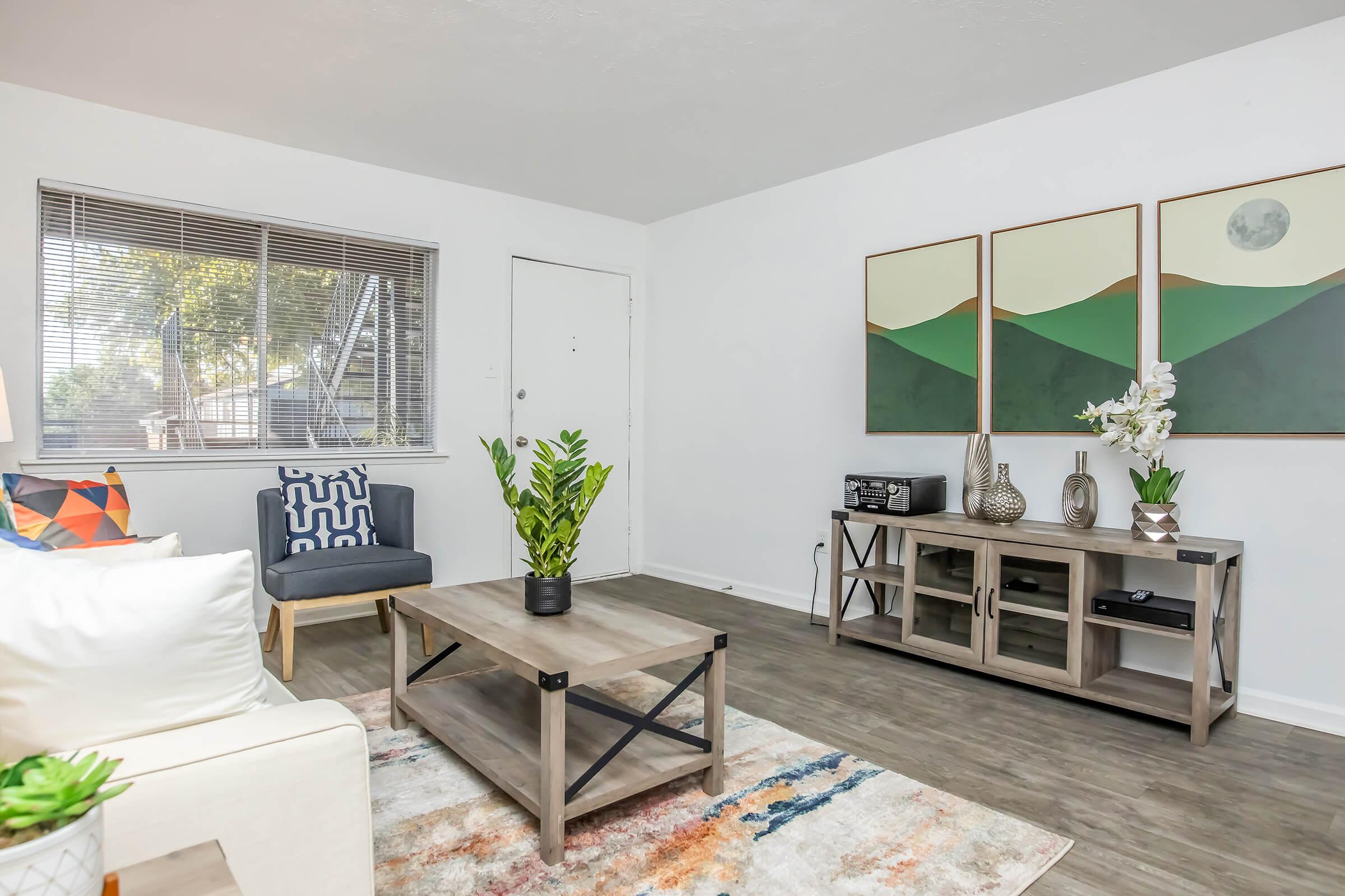 A bright and modern living room featuring a light-colored sofa, a colorful accent pillow, and a potted plant on a coffee table. A gray chair sits in the corner, and two framed landscape artworks adorn the wall. Natural light streams in through a window, illuminating the space with a warm ambiance.