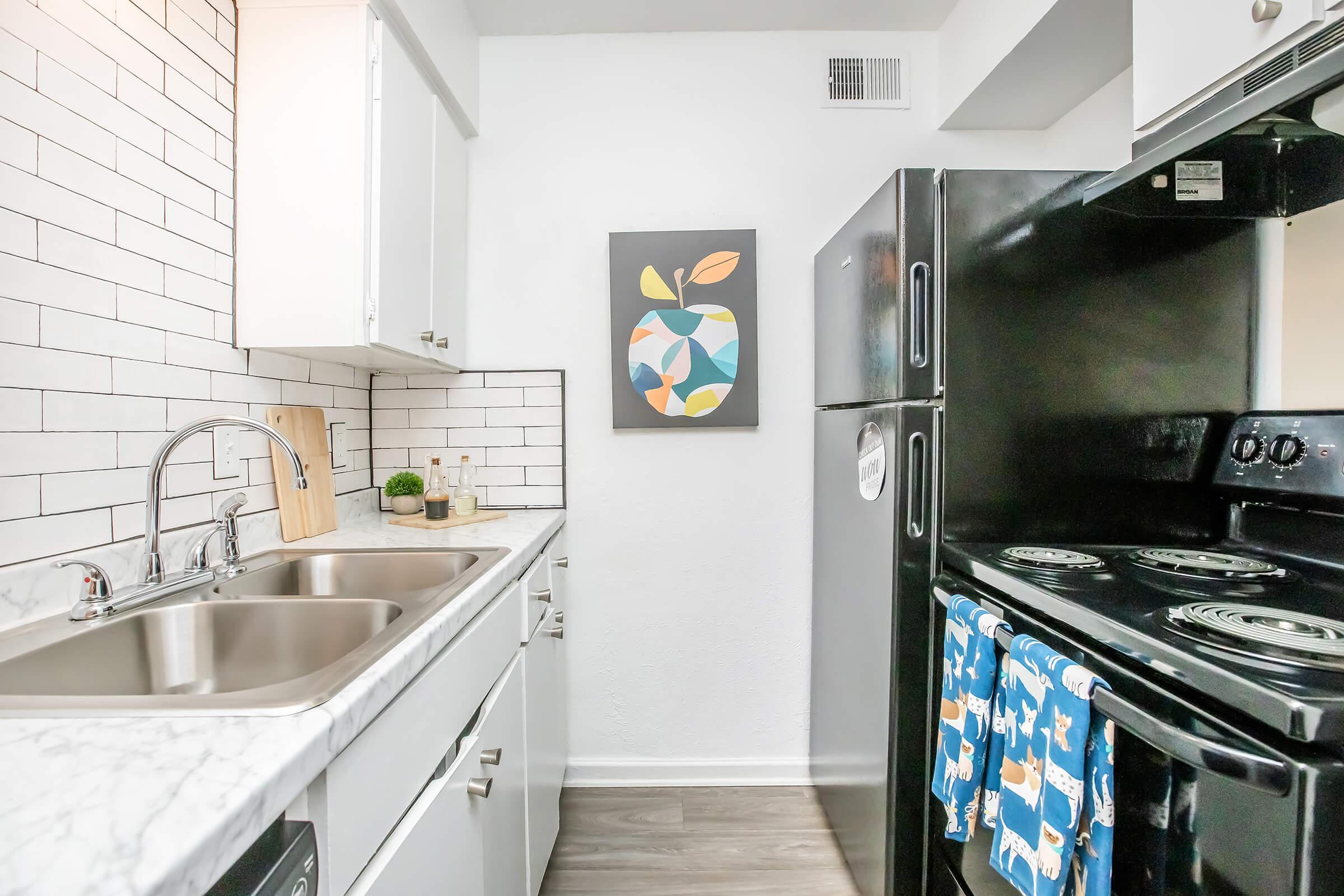 Modern kitchen with white cabinetry, a marble countertop, and a black stove with a blue kitchen towel. A black refrigerator stands nearby, and a colorful abstract fruit-themed artwork is displayed on the wall. The space features bright lighting and a clean, minimalist design.