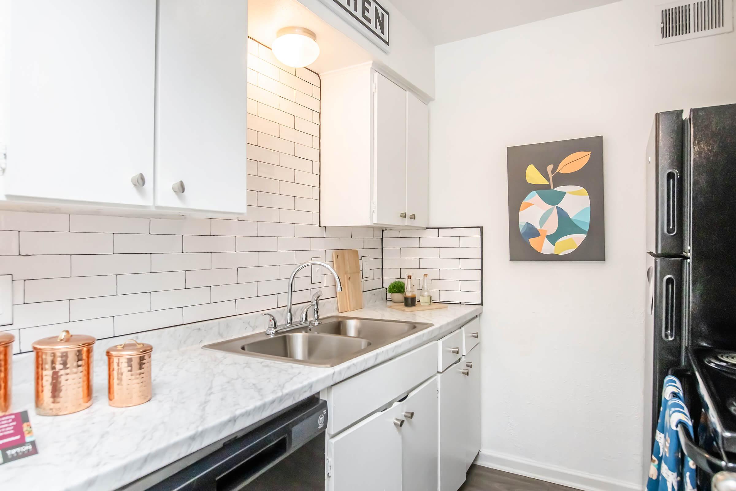 A modern kitchen featuring white cabinets, a double sink with a marble countertop, and a tiled backsplash. The kitchen includes copper canisters and a black refrigerator. A colorful abstract wall art piece is displayed above the countertop, adding a vibrant touch to the space.