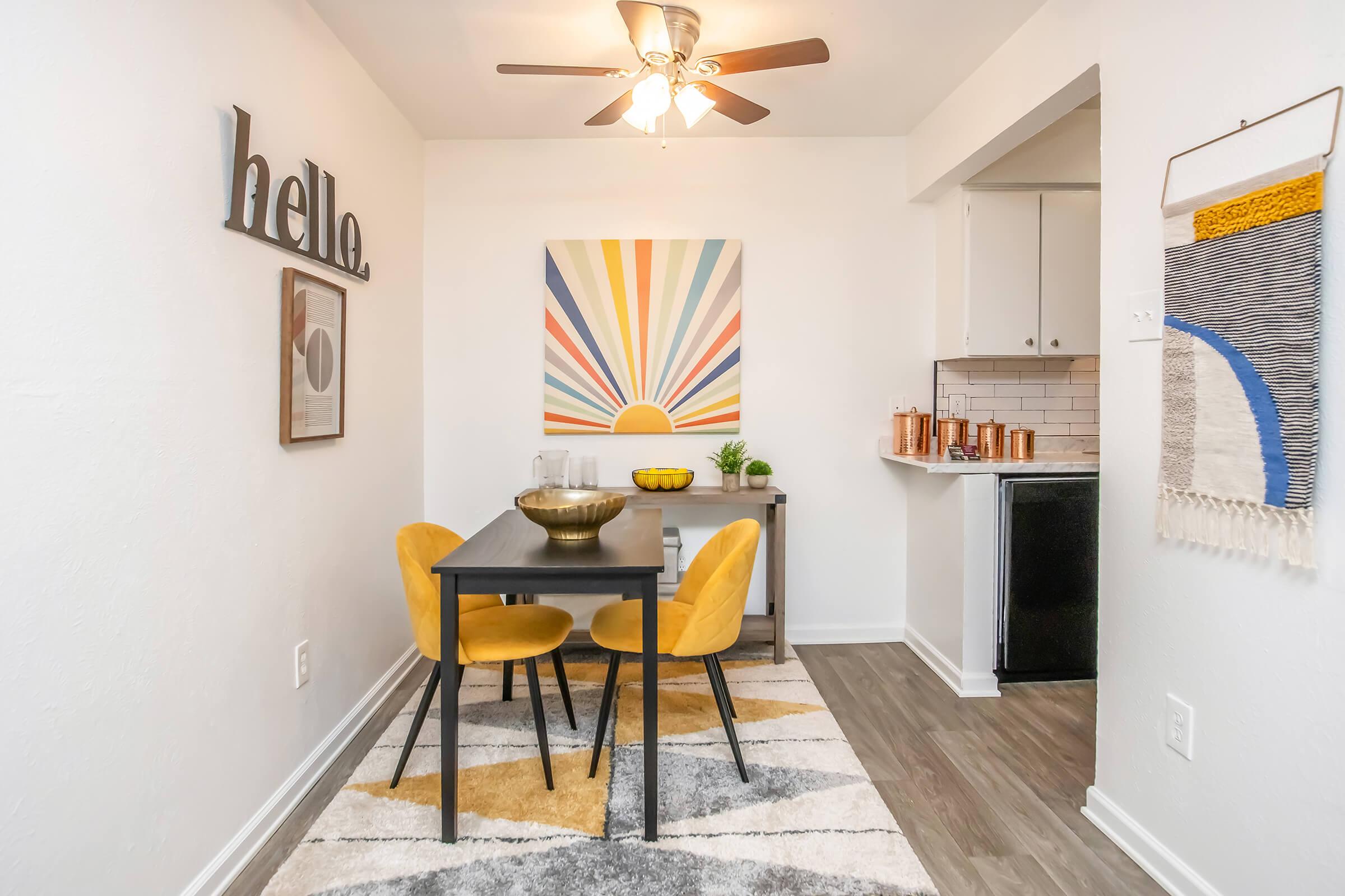 A cozy dining area featuring a black table with two yellow chairs. A colorful sunburst artwork hangs on the wall above, and a decorative bowl sits on the table. Soft lighting from a ceiling fan illuminates the space, which includes a patterned rug and a small kitchen area in the background.