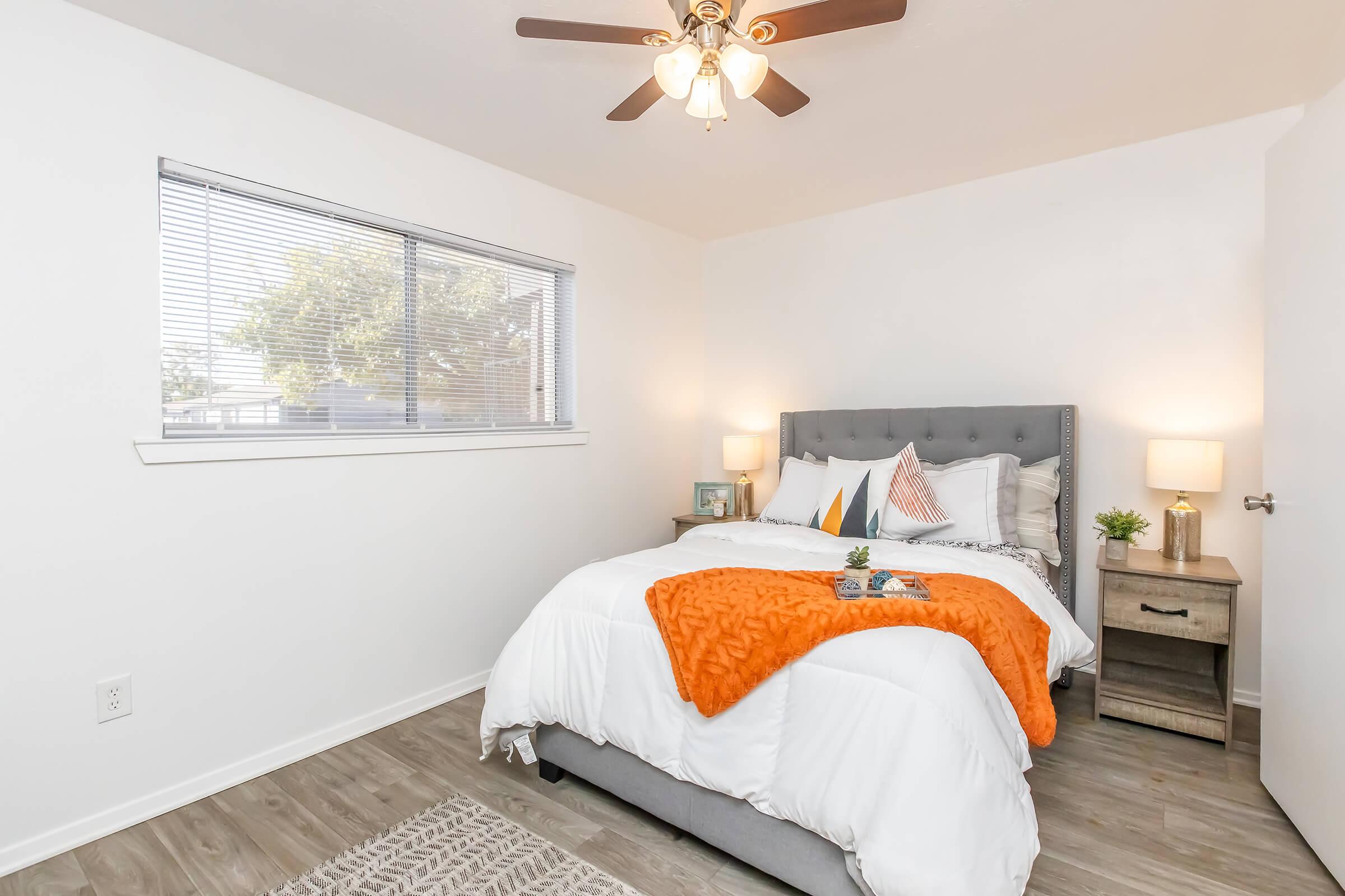 A cozy bedroom featuring a gray upholstered bed with white and orange bedding, two bedside tables with lamps, and a decorative plant. Natural light filters through a window with blinds, illuminating the room's modern decor and wooden flooring.