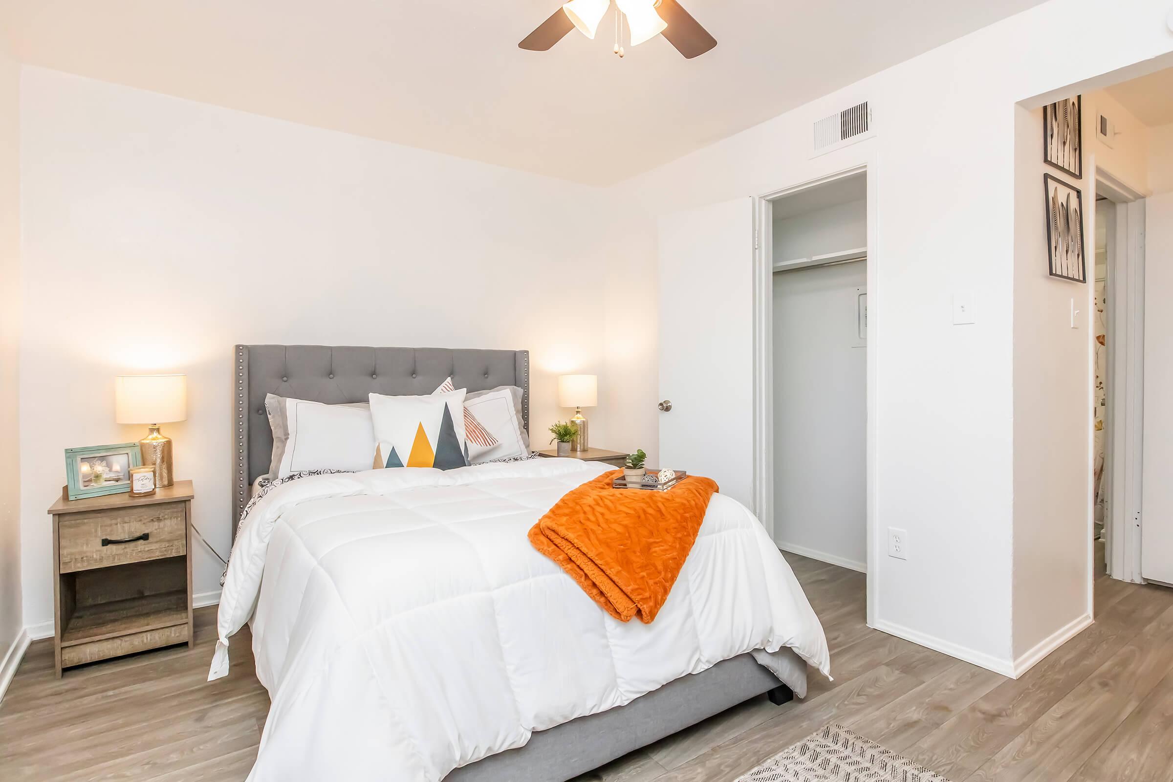 A cozy bedroom featuring a gray upholstered bed with white bedding and decorative pillows in orange and blue. There are two nightstands with lamps, a small plant, and framed pictures on the walls. A closet is visible, and the flooring is light wood.