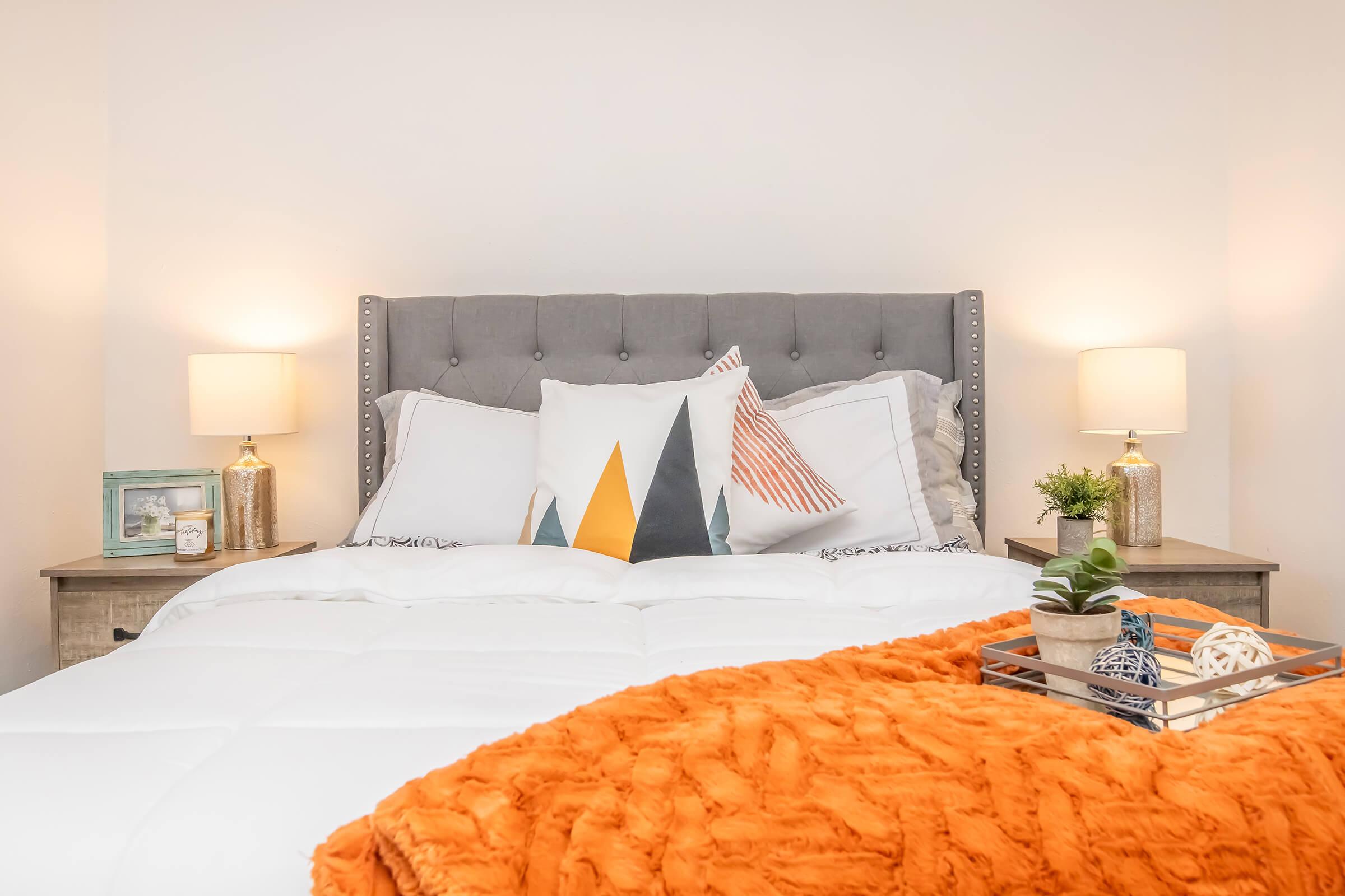A stylish bedroom featuring a gray upholstered headboard, a neatly made bed with white linens and an orange throw blanket. There are decorative pillows with geometric patterns, bedside tables with white lamps, and a small plant on a tray. The walls are light-toned, creating a cozy atmosphere.
