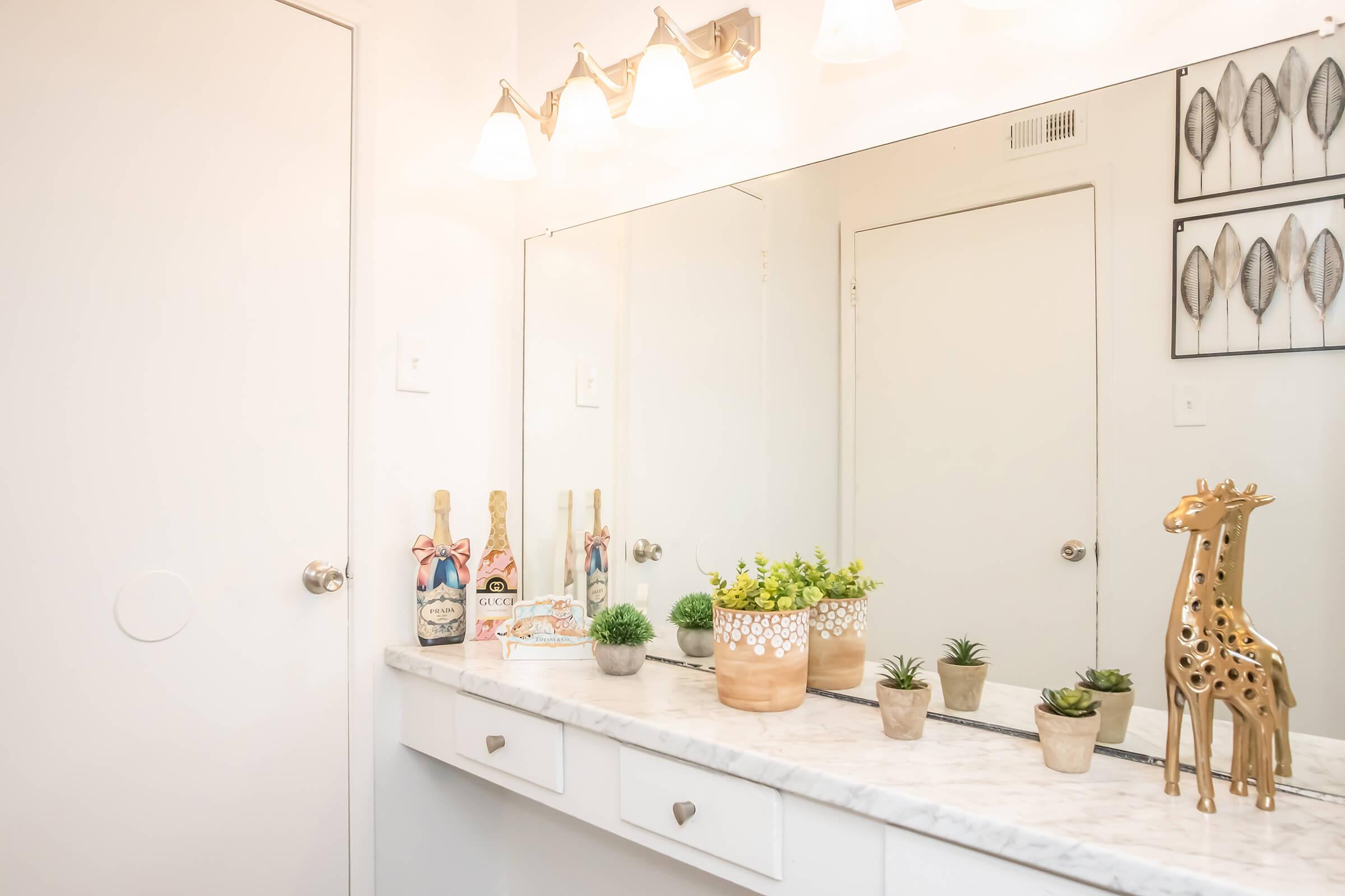 A well-lit bathroom with a marble countertop. The mirror reflects the space, and there are decorative items including potted plants, a golden giraffe figurine, and small ornaments. Soft lighting from above adds warmth to the minimalist decor. A doorway is visible in the background.