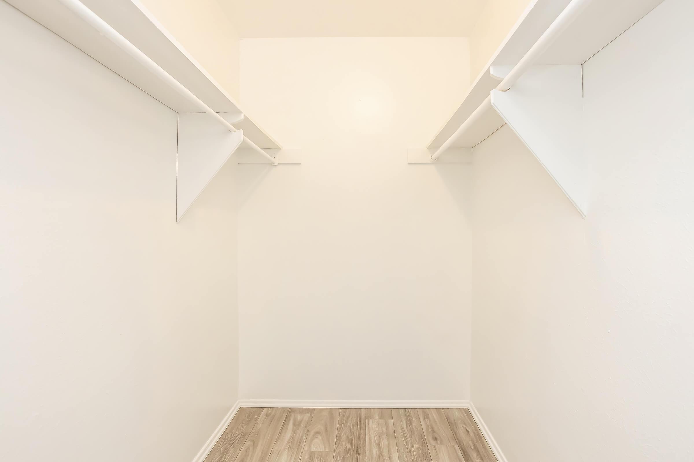 Empty closet with light-colored walls and wooden flooring. Two shelves on either side, creating an open space in the center. The overall atmosphere is clean and minimalistic.