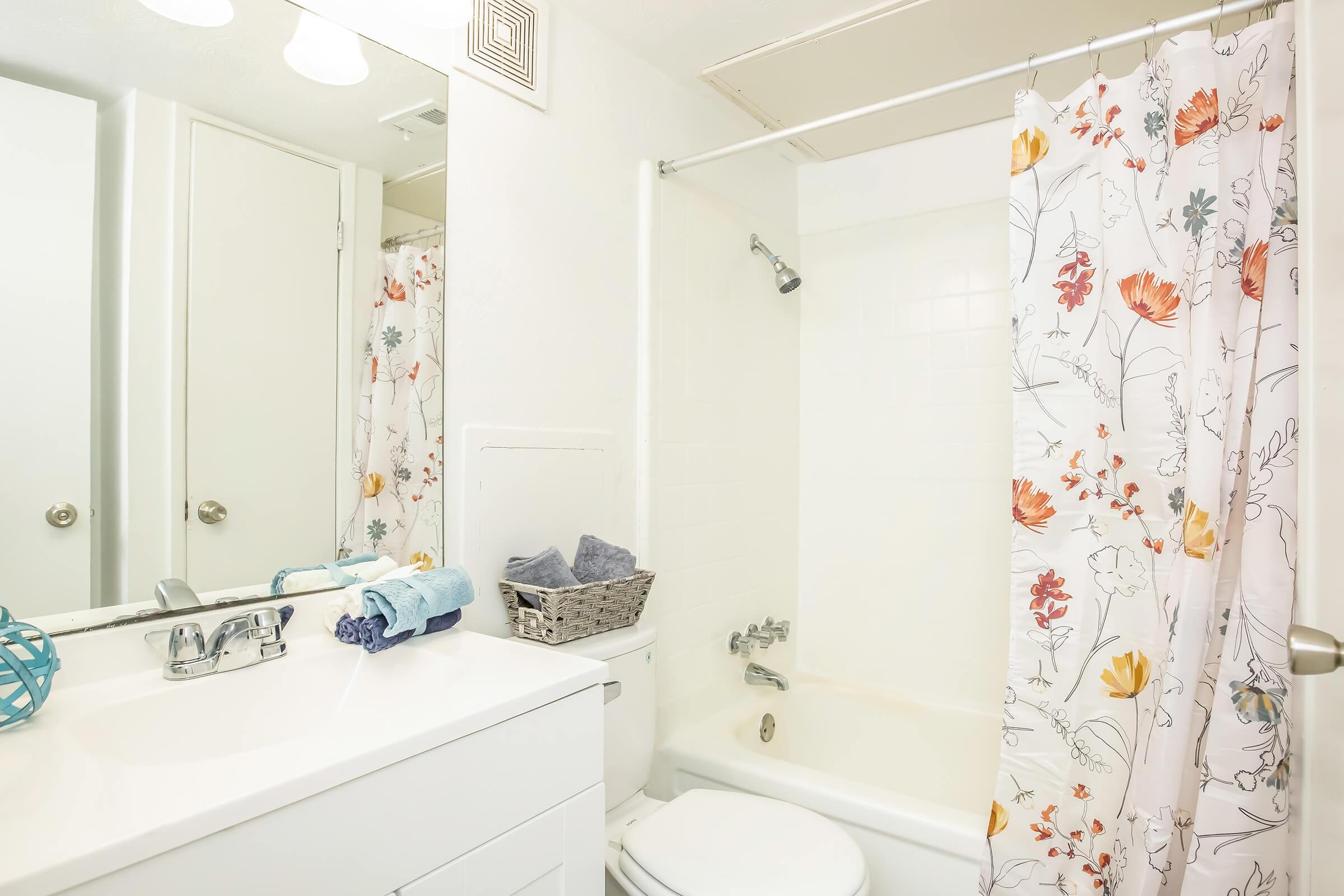 A bright bathroom featuring a white sink and cabinet, a bathtub with a shower, and a floral shower curtain. There's a woven basket with towels on the edge of the tub, and decorative items on the countertop, creating a clean and inviting space.